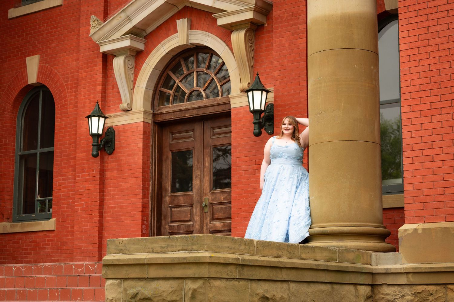 Woman in light blue ball gown posing on steps of red brick historic building.