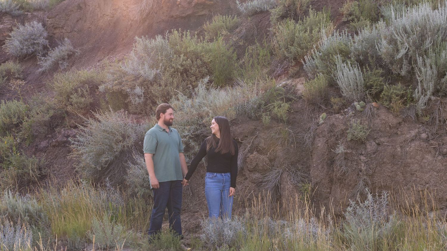 Couple standing together in a scenic landscape with sagebrush and rocky hillside backdrop.