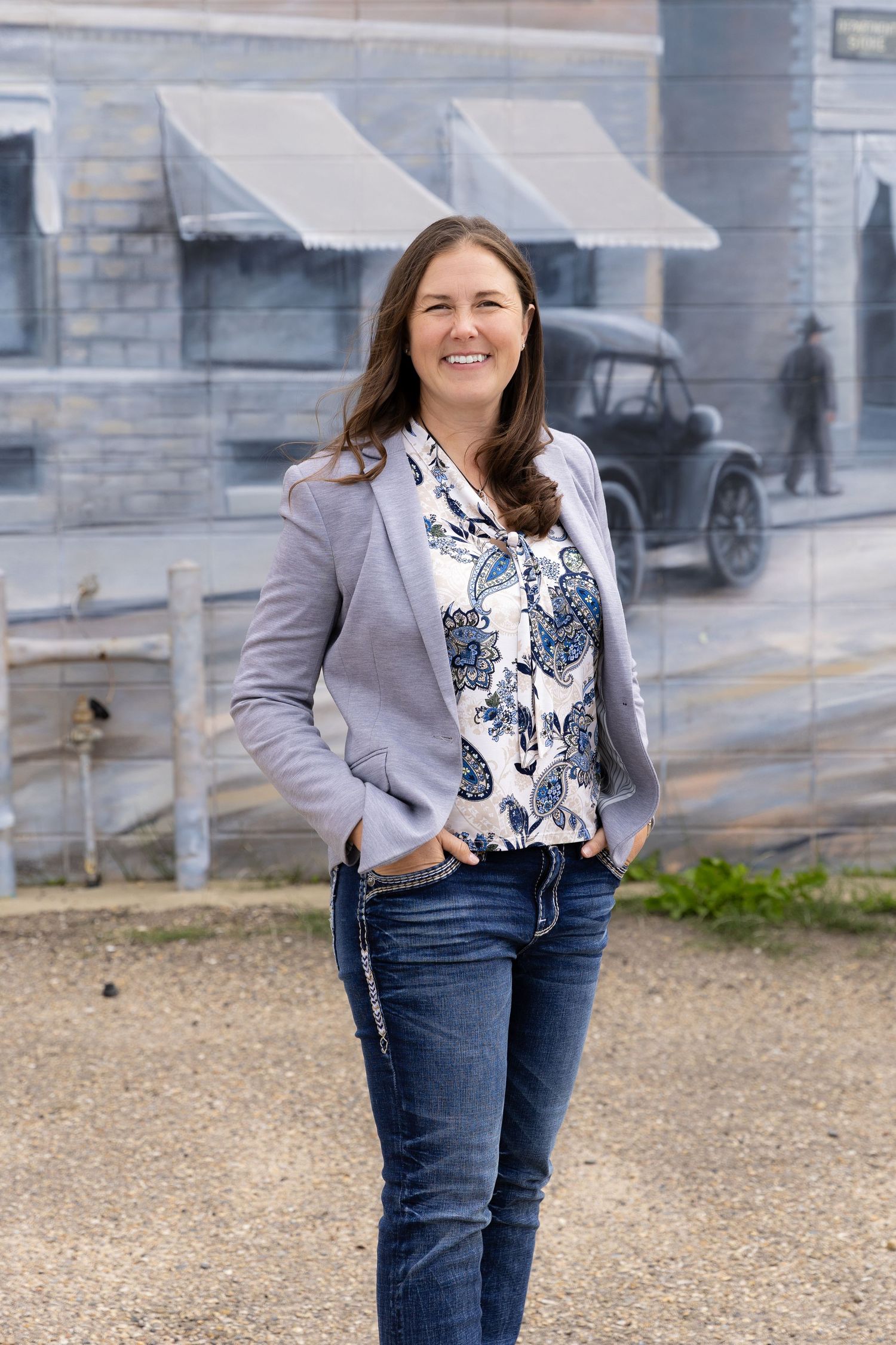 Woman in gray blazer and floral top smiling in front of a vintage street mural outdoors.