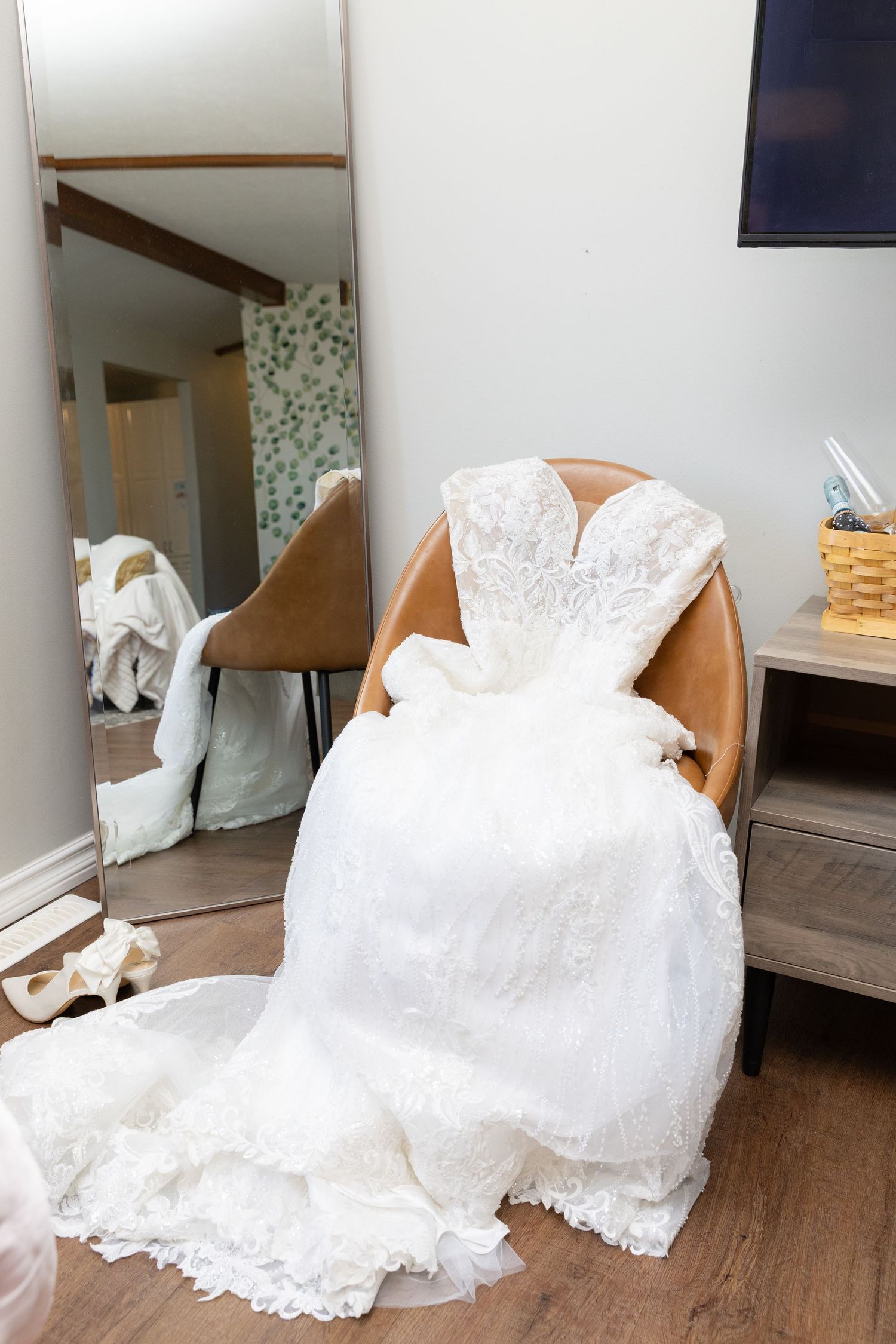 White lace wedding gown draped over a leather chair beside a full-length mirror in a bridal suite.