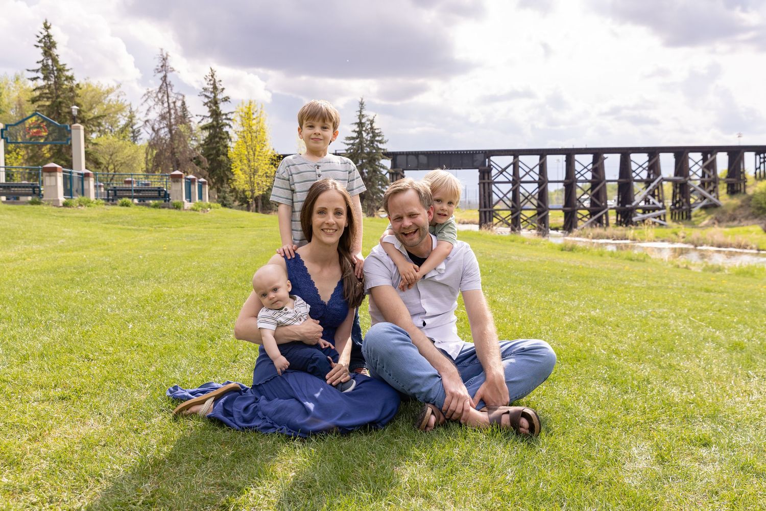 Family of four sitting on green grass outdoors near a wooden trestle bridge on a cloudy day.