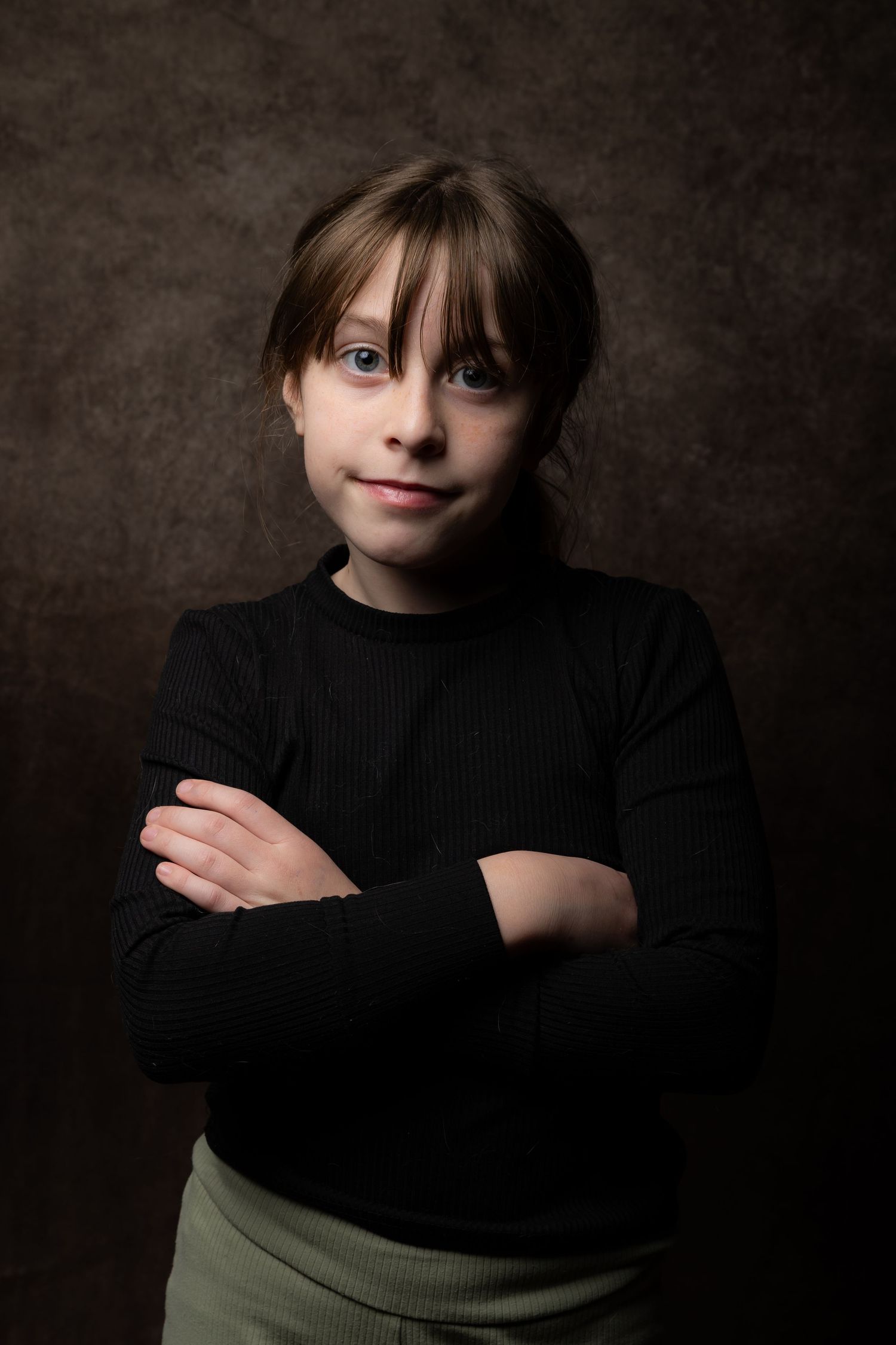 Young girl with blue eyes in a black turtleneck sweater posing with arms crossed on dark background.