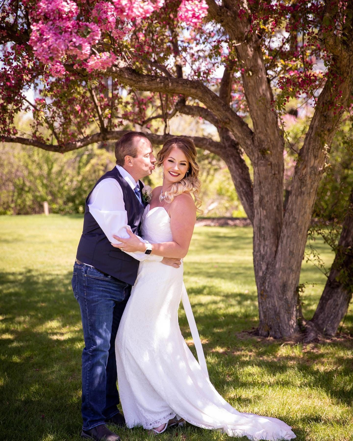 Smiling bride and groom embracing under a stunning pink flowering tree in a lush green garden setting.