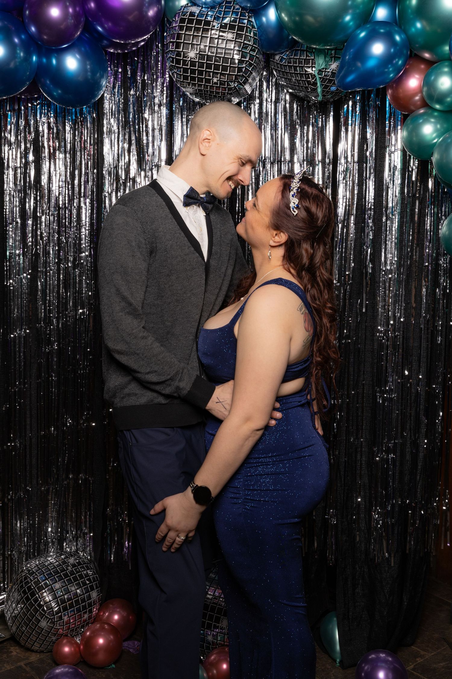 Couple in formal attire smiling face-to-face in front of a glittery backdrop with colorful balloons.