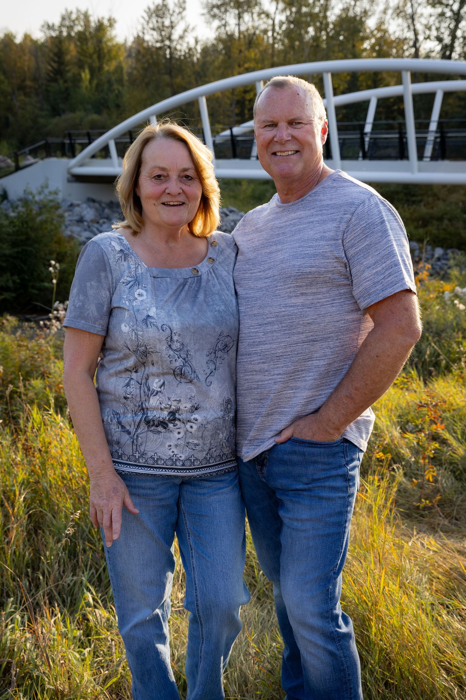 Smiling mature couple standing together in a golden field with a white arch bridge behind them.
