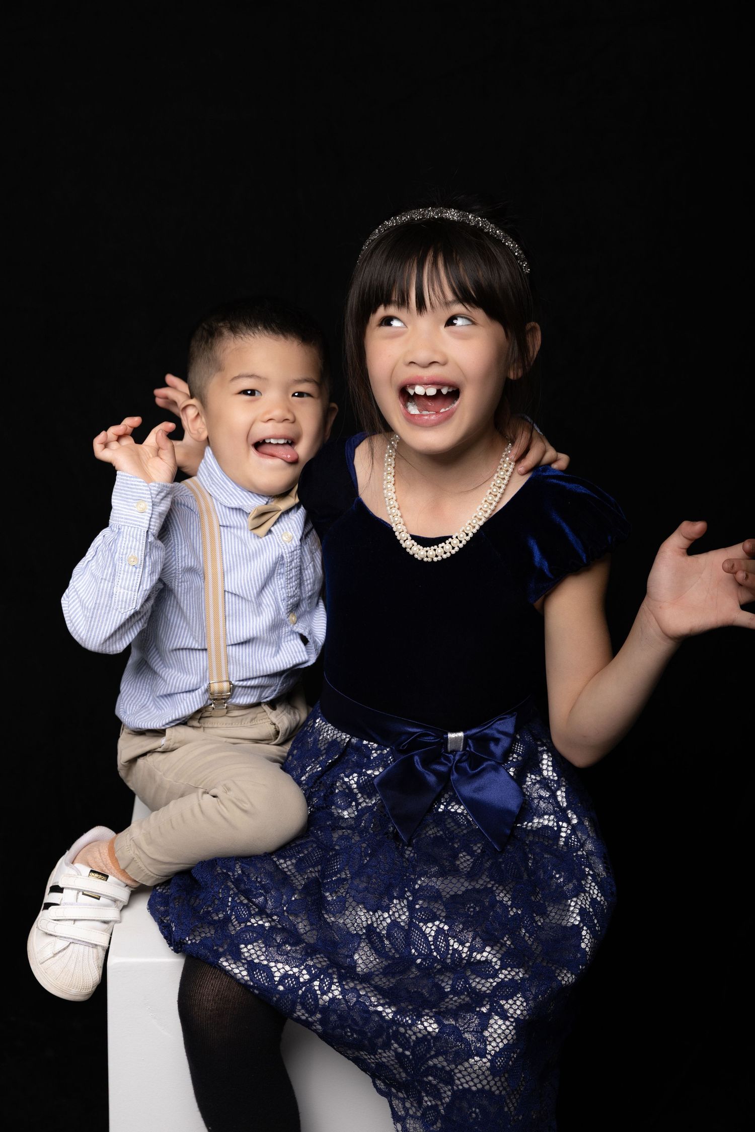 Two young siblings laughing and playing together in formal dress against black background.