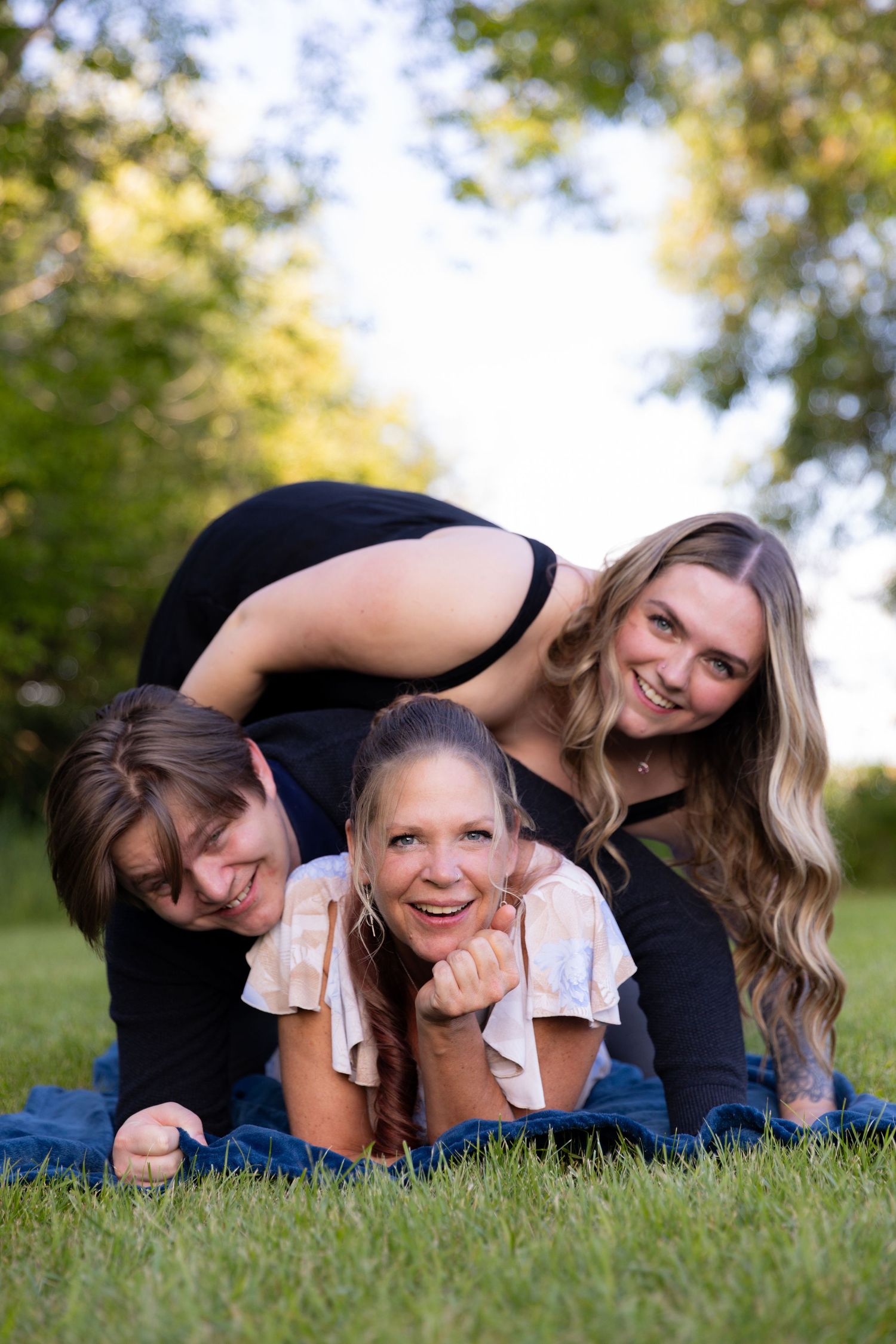 Family of 3 laughing and stacking on top of each other while lying on green grass outdoors.