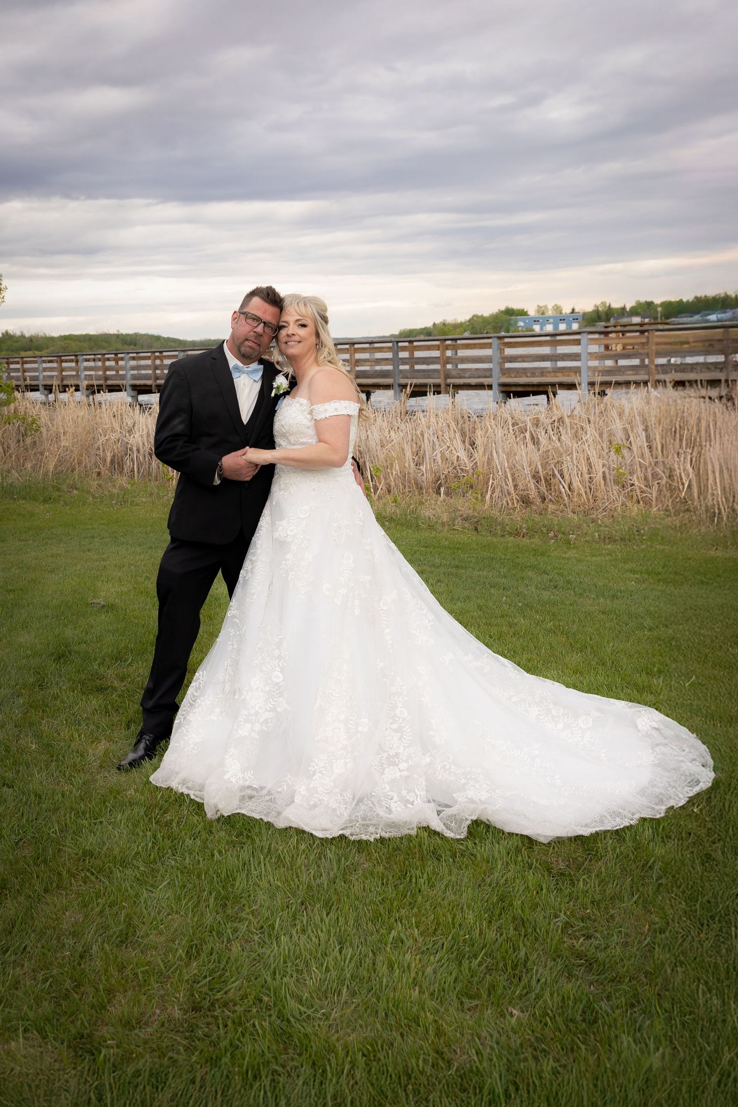 Bride and groom posing outdoors on green grass with tall reeds and a wooden bridge in the background on their wedding day.