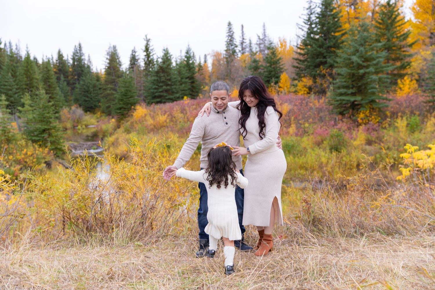 A joyful family plays together in a beautiful fall setting surrounded by colorful autumn foliage and evergreen trees.