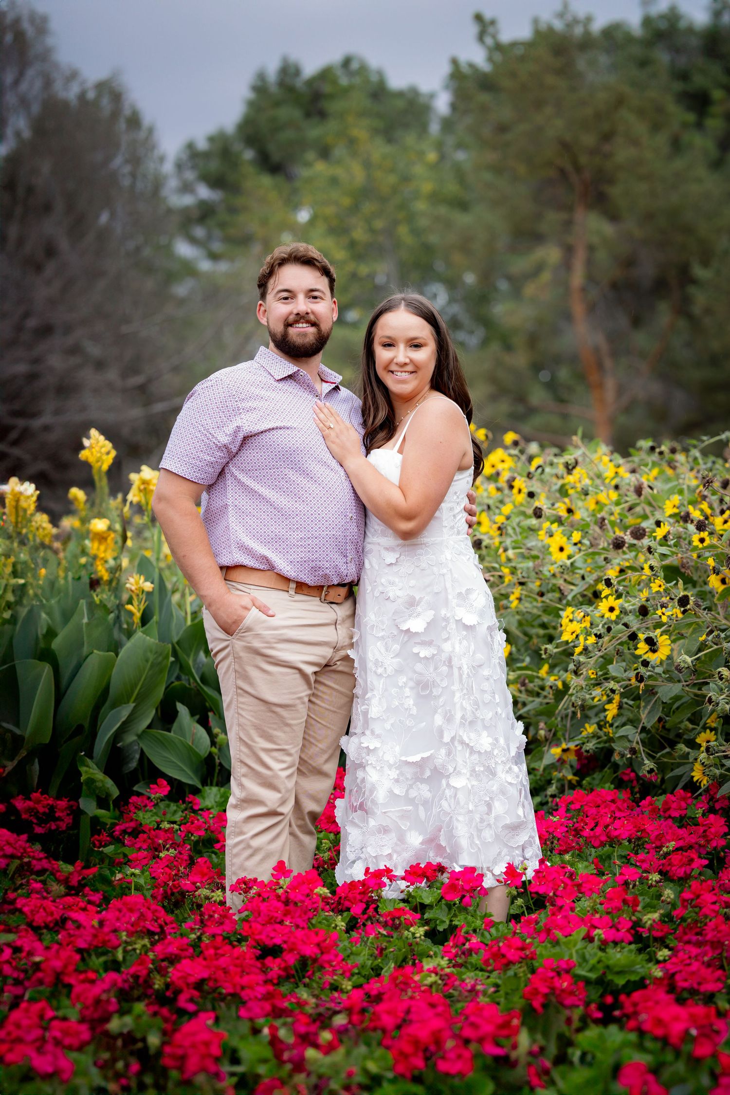 Smiling couple posing in a vibrant garden with red and yellow flowers, woman in white dress.