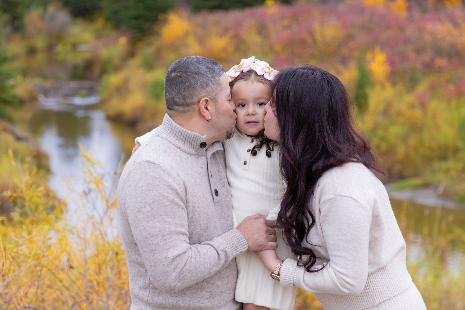 Parents kissing their young daughter on cheeks outdoors with fall foliage backdrop.