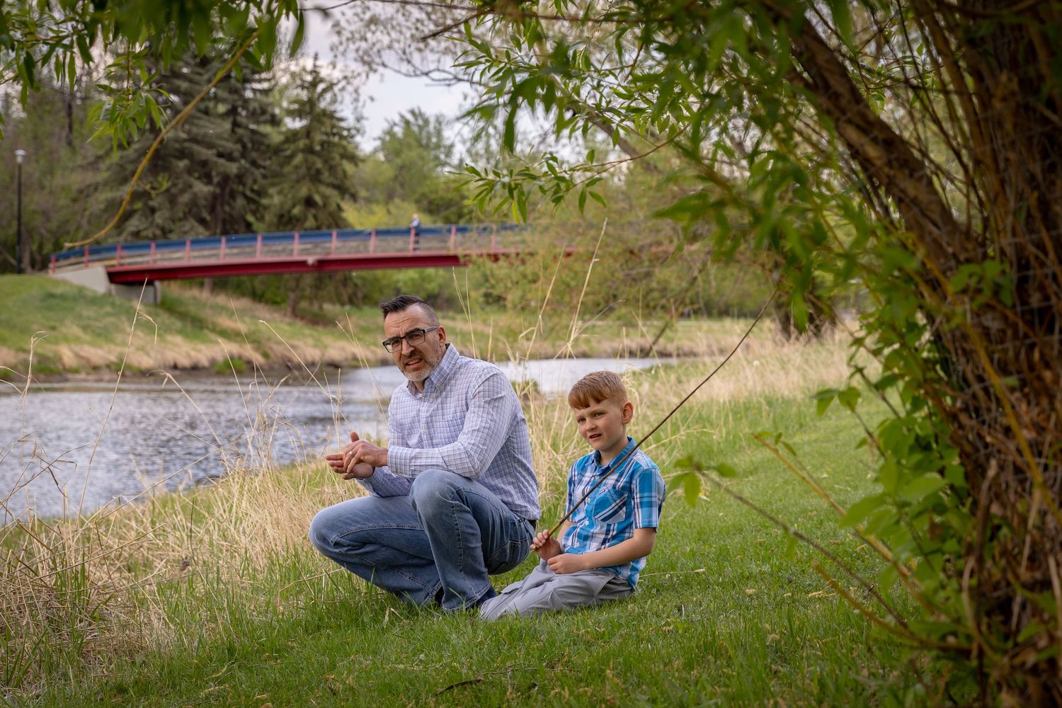 Father and young son sitting together near a river fishing under a tree with a red bridge behind.