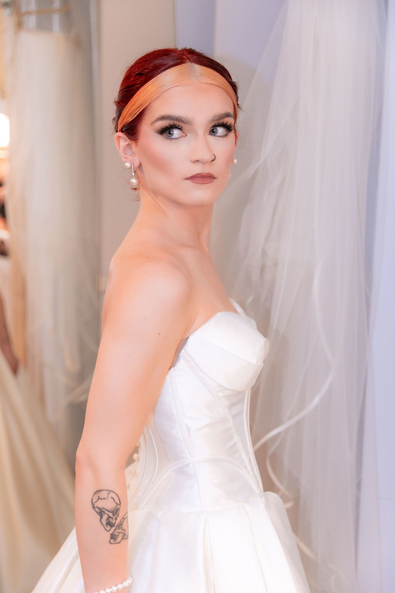 Beautiful red-haired bride in a strapless white ball gown with arm tattoo posing elegantly in a bridal boutique.