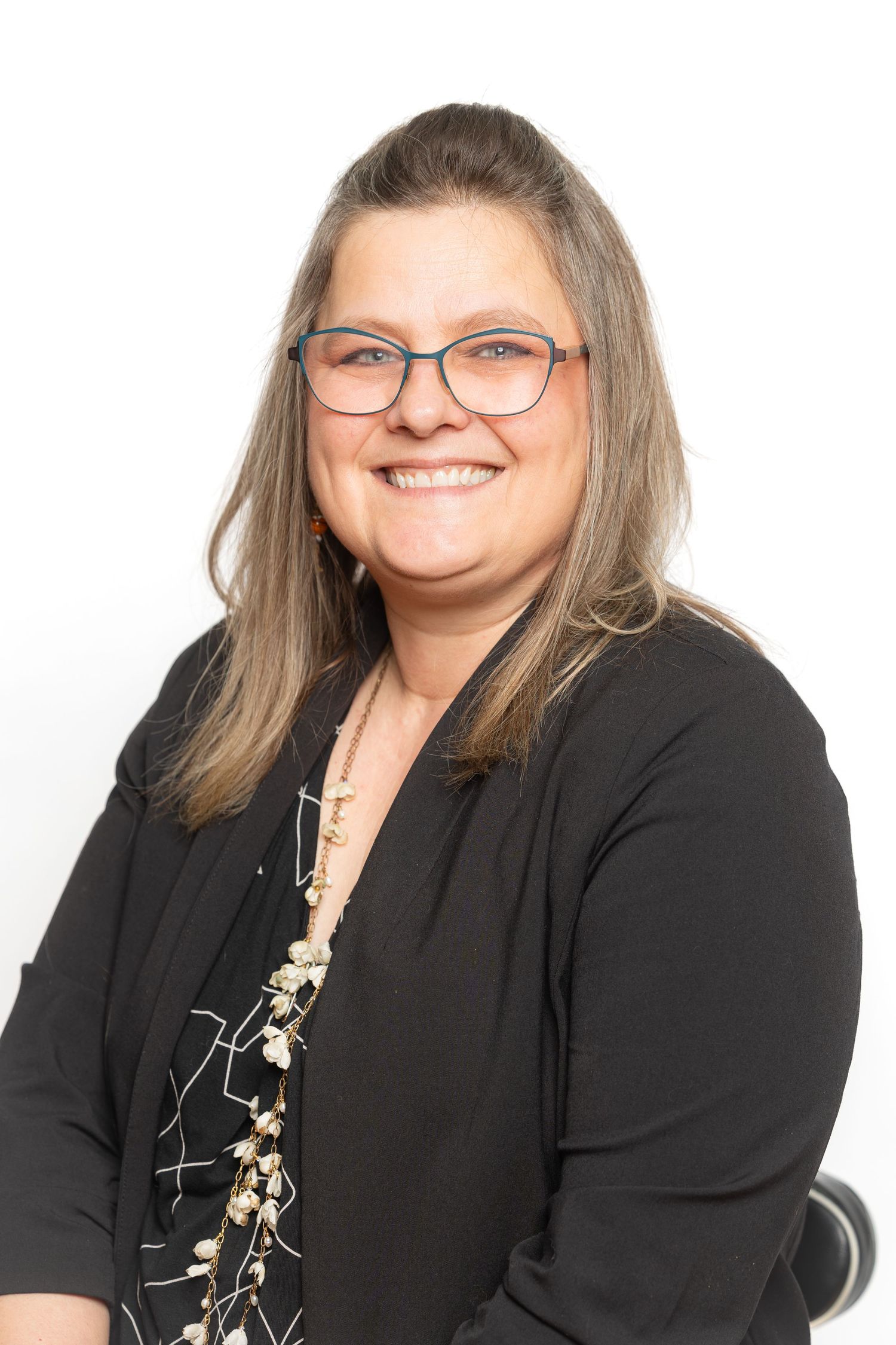 Smiling woman with glasses and gray-streaked hair wearing a black blazer and floral blouse on white background.