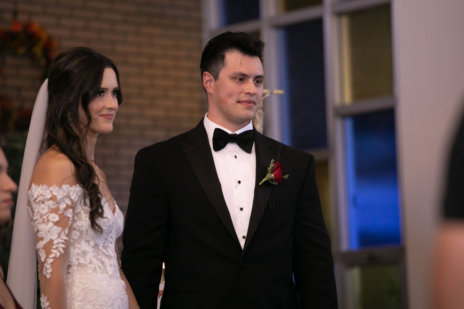 Bride and groom standing at the altar during their wedding ceremony, groom wearing a black tuxedo with a red boutonniere.
