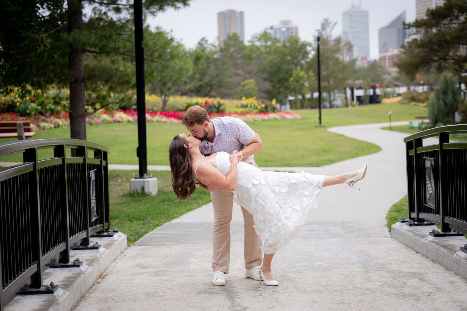 Couple sharing a romantic dip kiss on a park pathway with city skyline in background.
