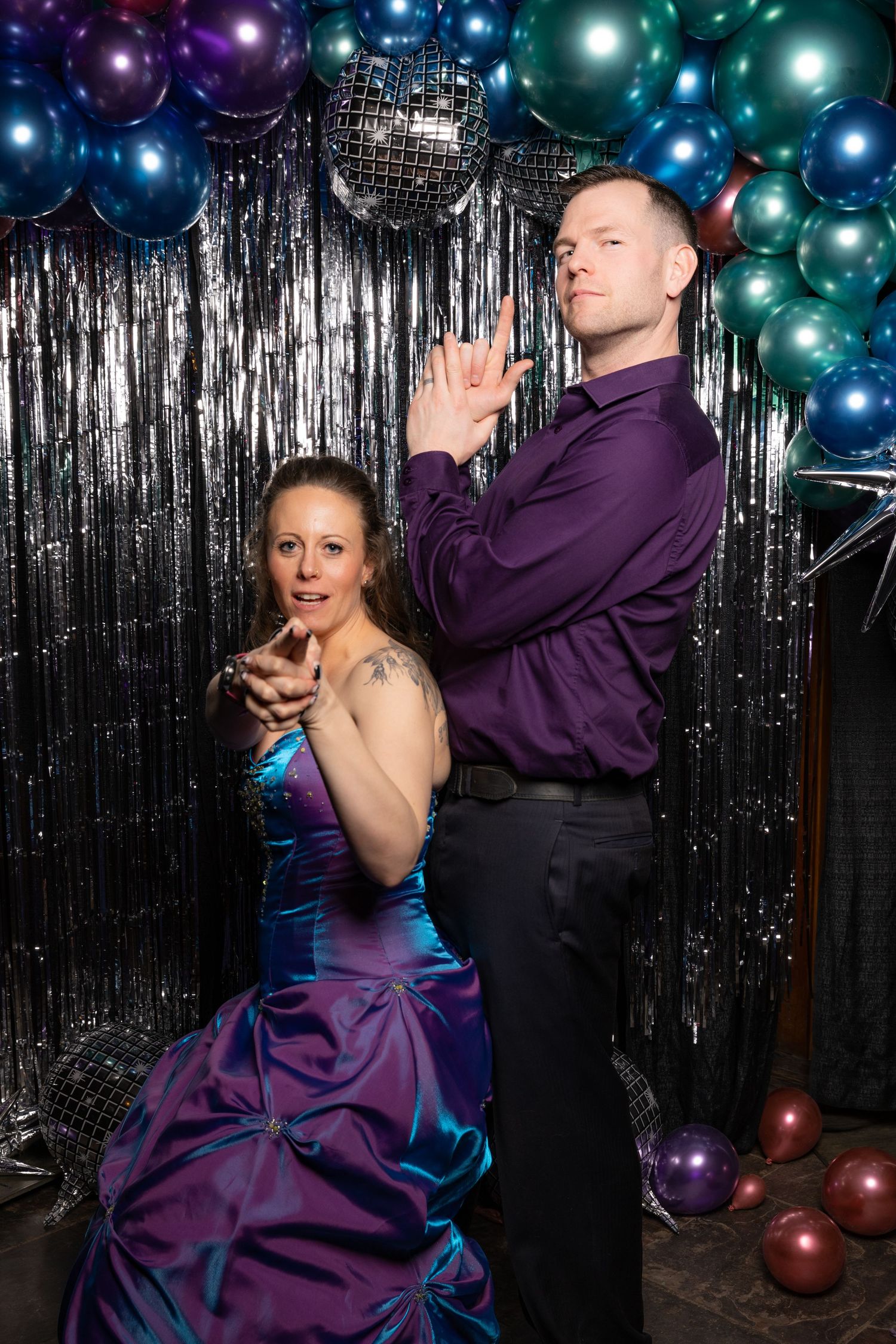 A couple poses dramatically at a prom-themed photo booth with colorful balloons and silver tinsel backdrop.
