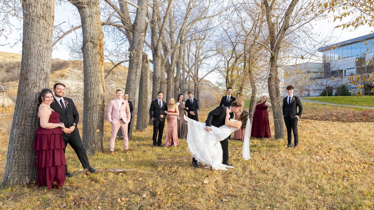 Wedding party group posing among trees in autumn landscape with bride and groom dip kiss centerpiece.