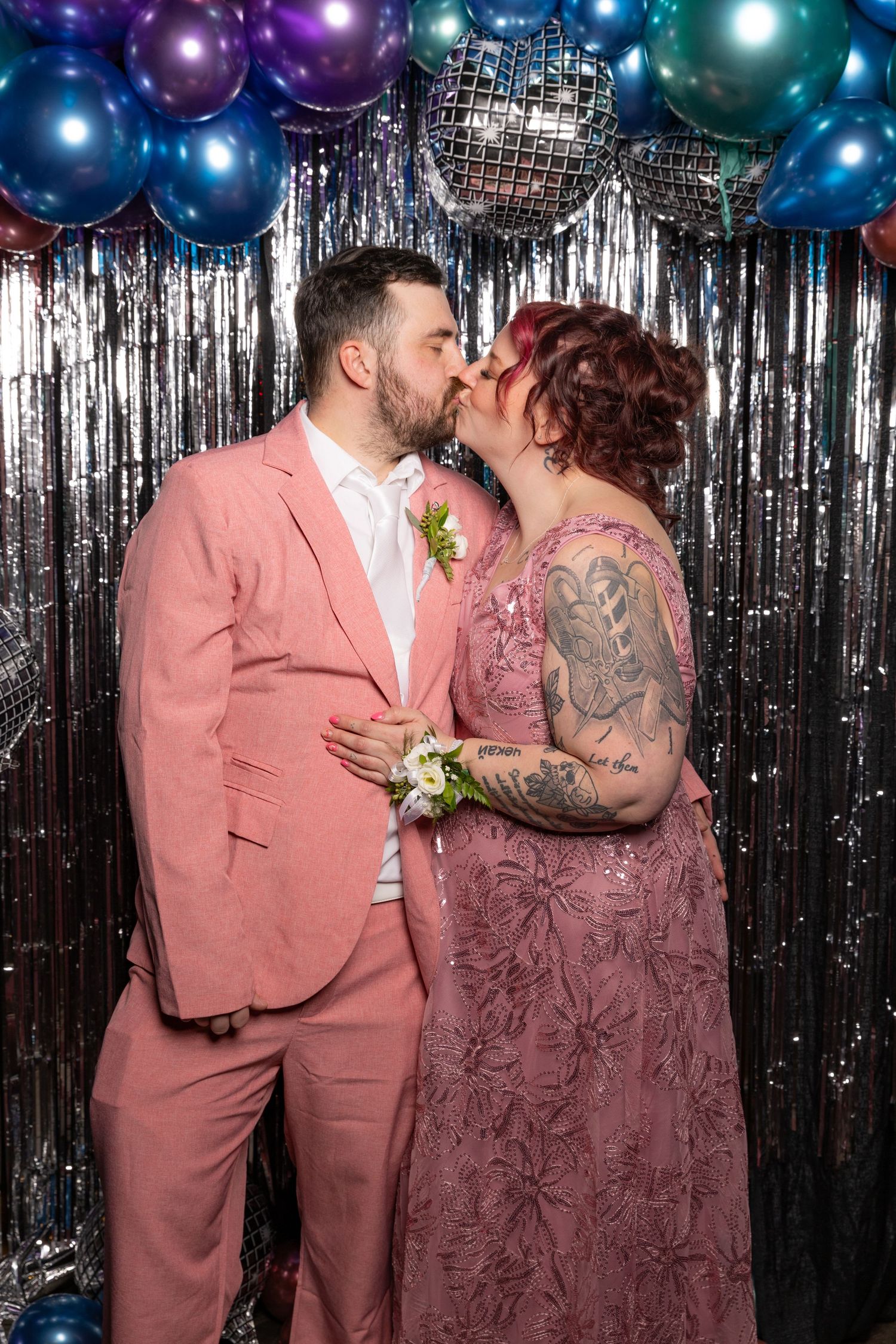 Couple kissing at prom, man in pink suit and woman in pink floral dress, balloon backdrop.