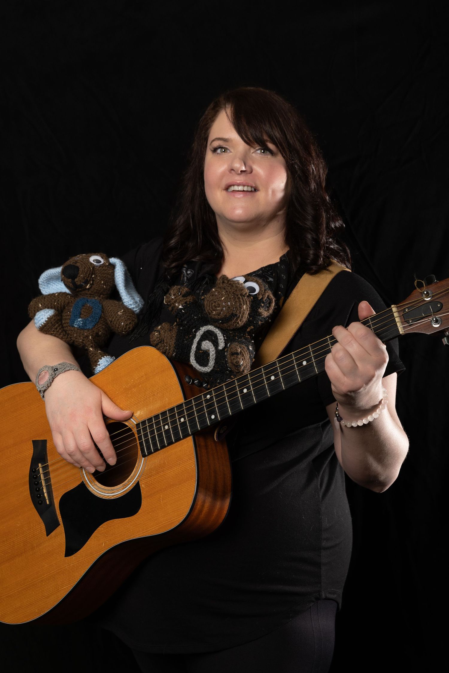 Woman in black playing acoustic guitar with teddy bears, smiling against dark background.
