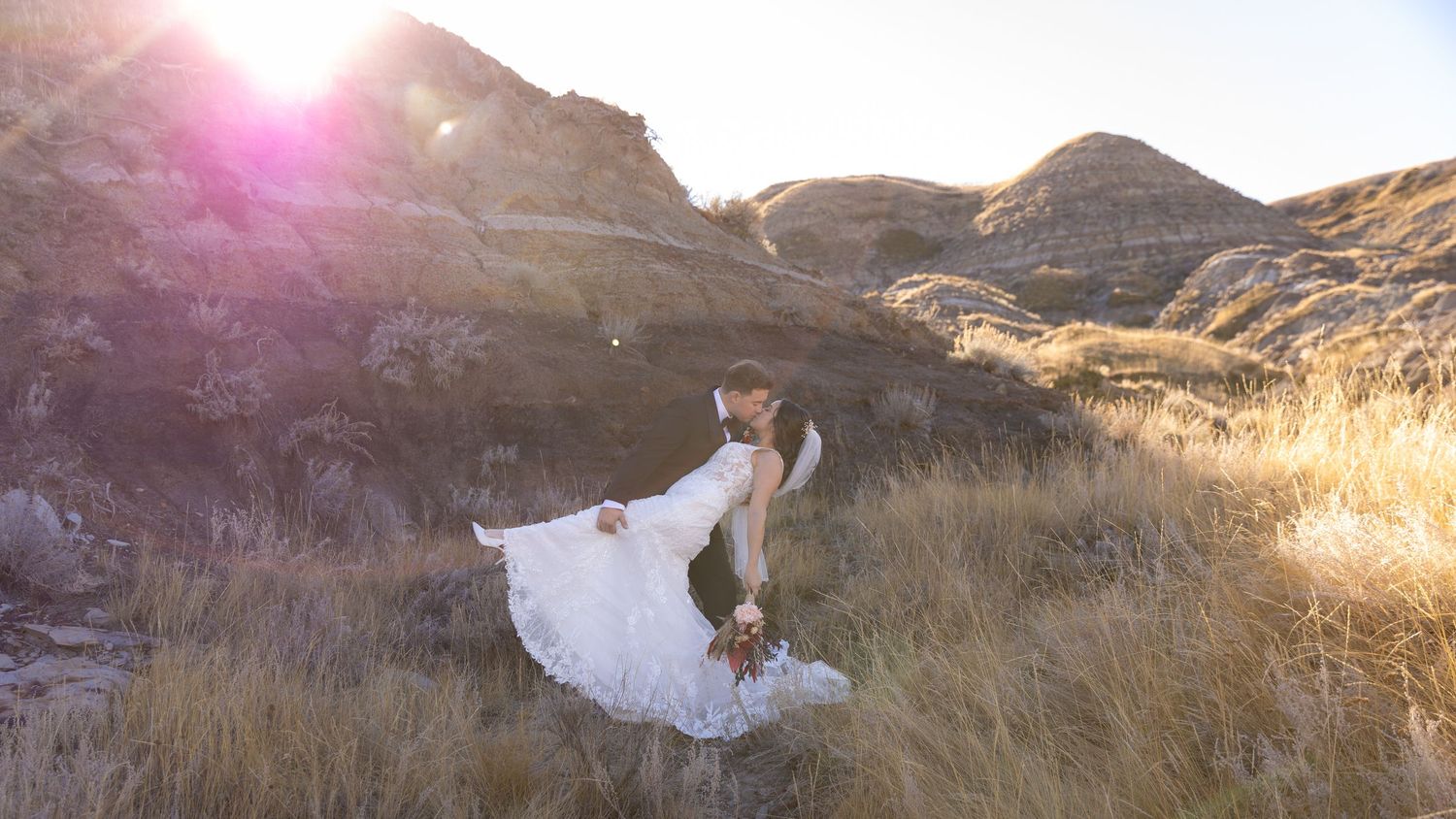 Bride and groom embracing in a dramatic sunlit landscape with rugged badlands hills in background.