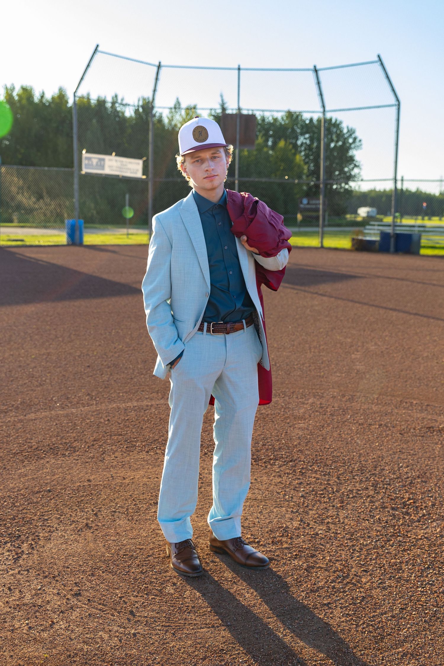 Young man in a light blue suit and white cap standing on a baseball diamond holding a maroon jacket.