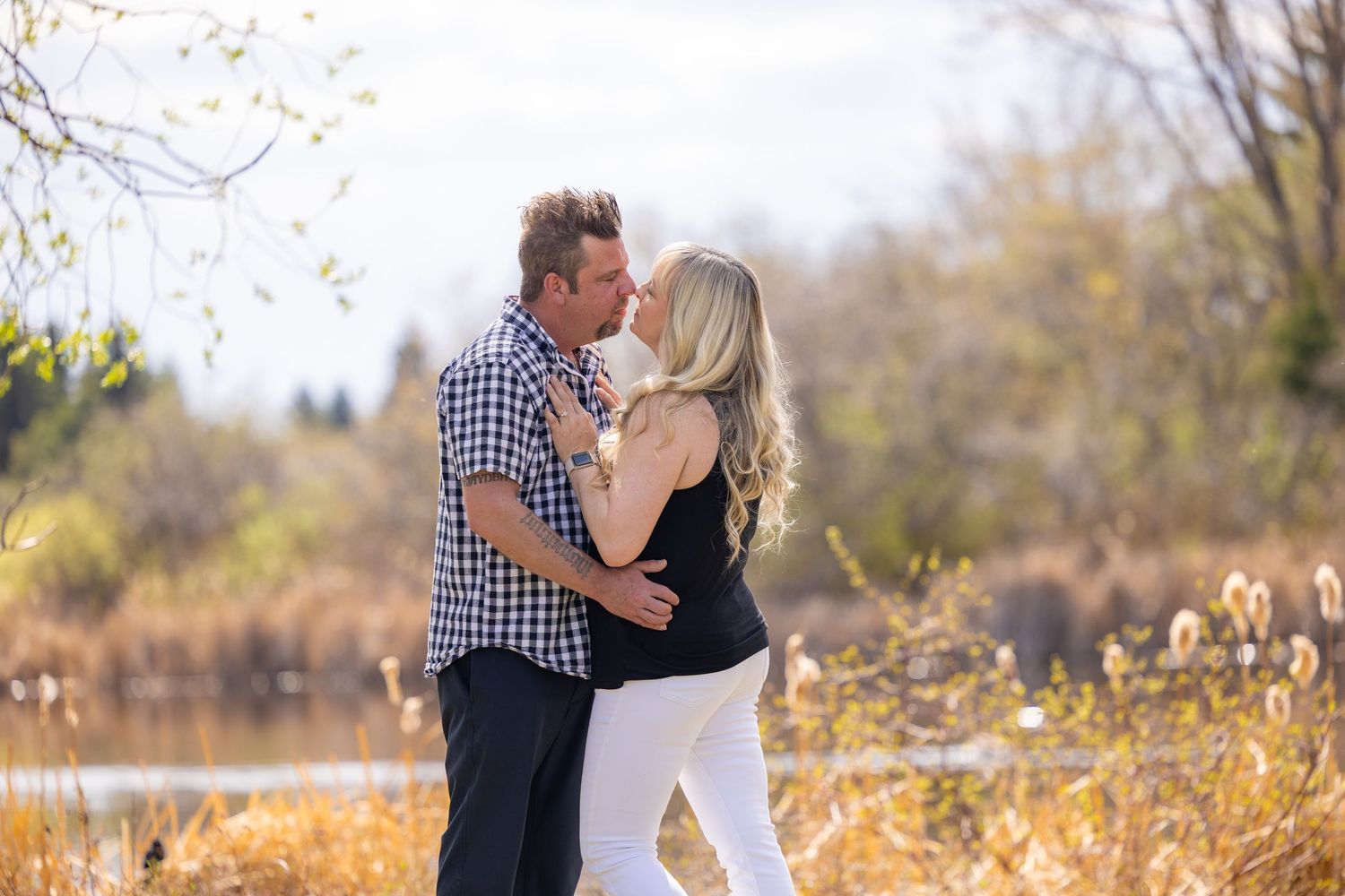 Romantic couple sharing a kiss outdoors near a pond with golden autumn foliage in the background. Leduc