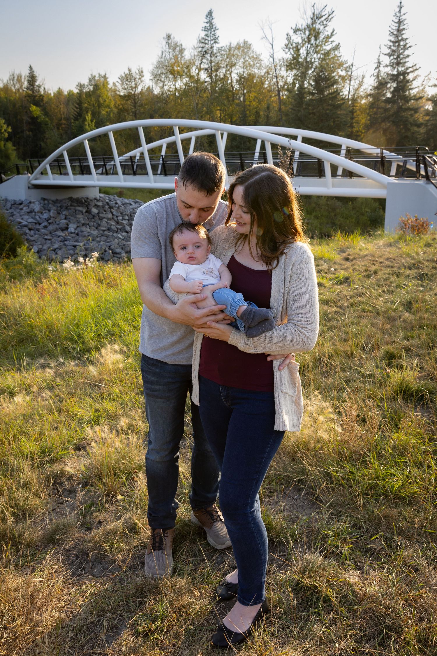 Young couple holding their newborn baby outdoors near a white arch bridge in golden hour sunlight.