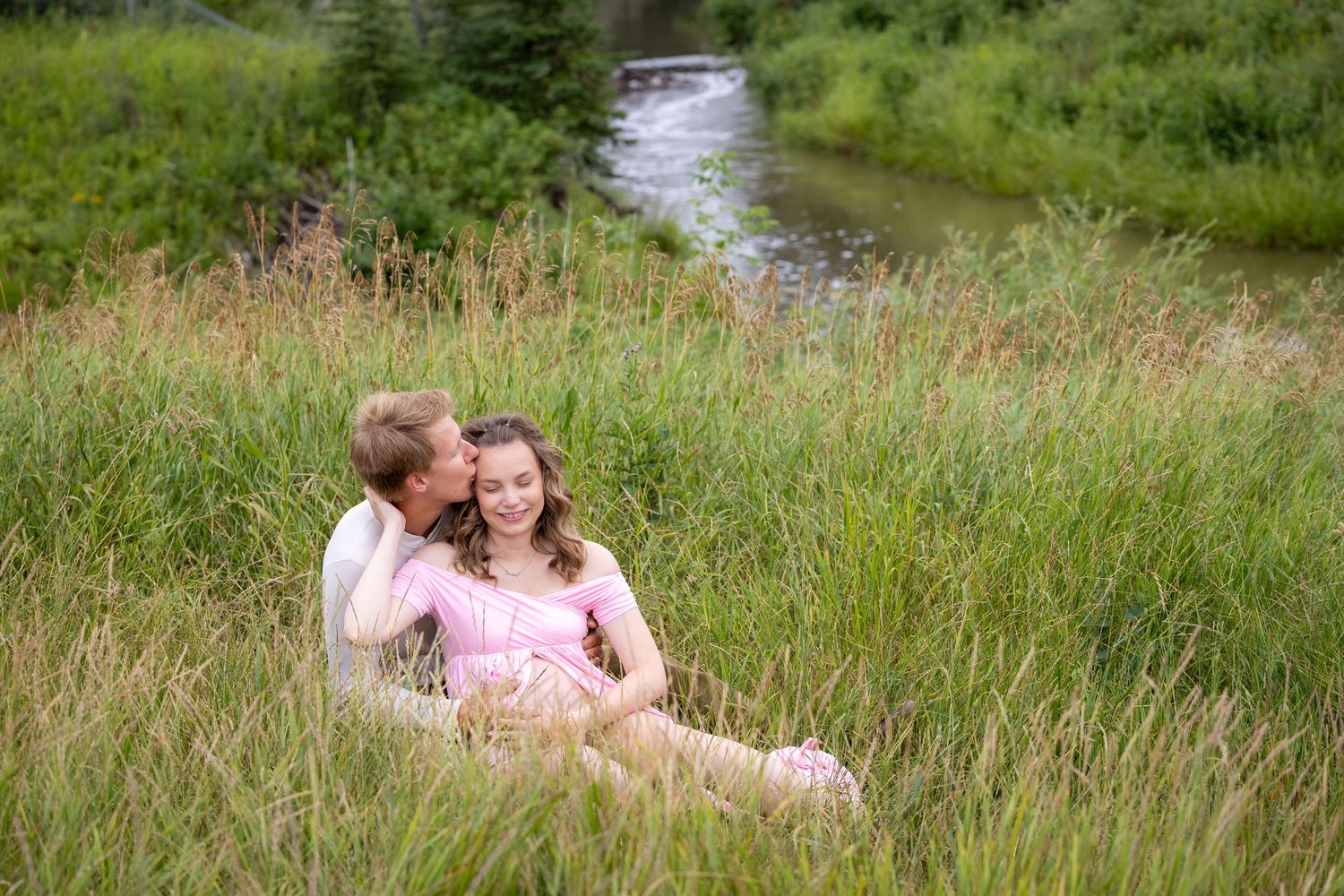 Couple sitting in tall grass by a stream, man kissing woman's cheek, woman in pink maternity dress.