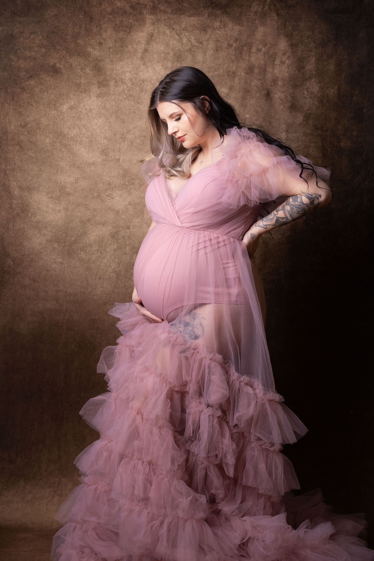 Pregnant woman in a flowing pink ruffled tulle maternity gown posing against a warm brown backdrop.