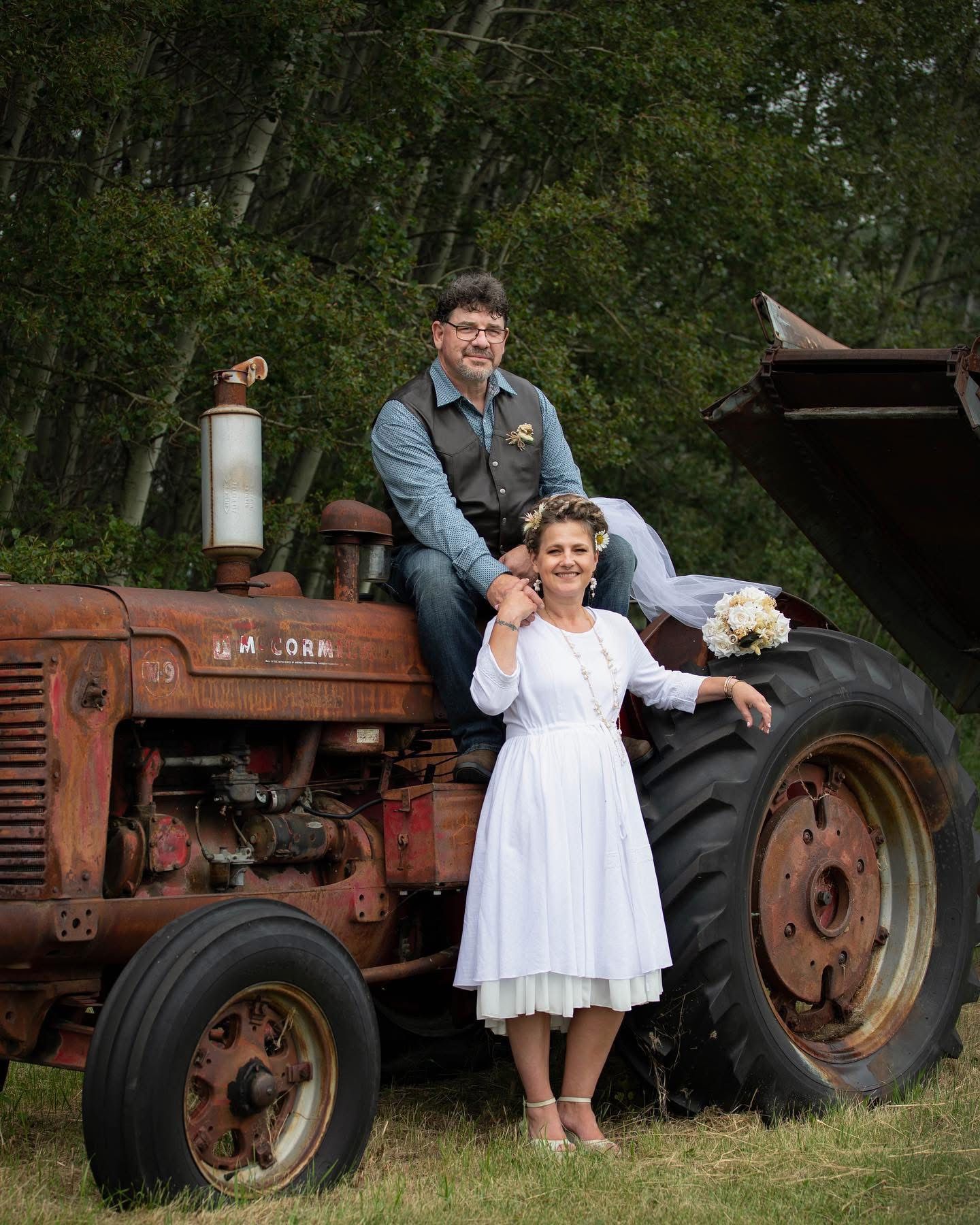 Bride in vintage white dress standing beside an old red tractor with groom seated on top in a rustic outdoor setting.