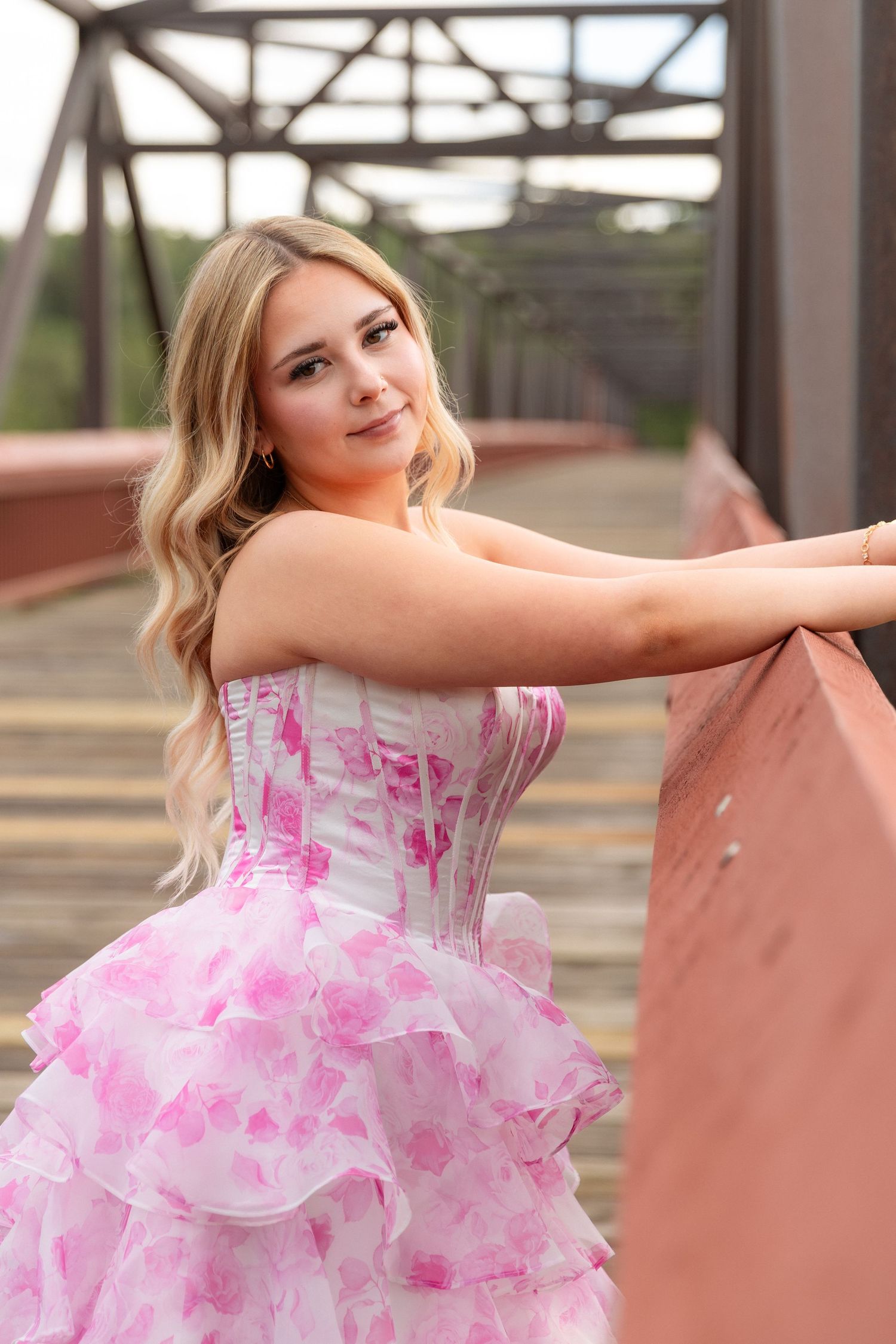 Young blonde woman in pink floral ruffled prom dress posing on a rustic bridge.