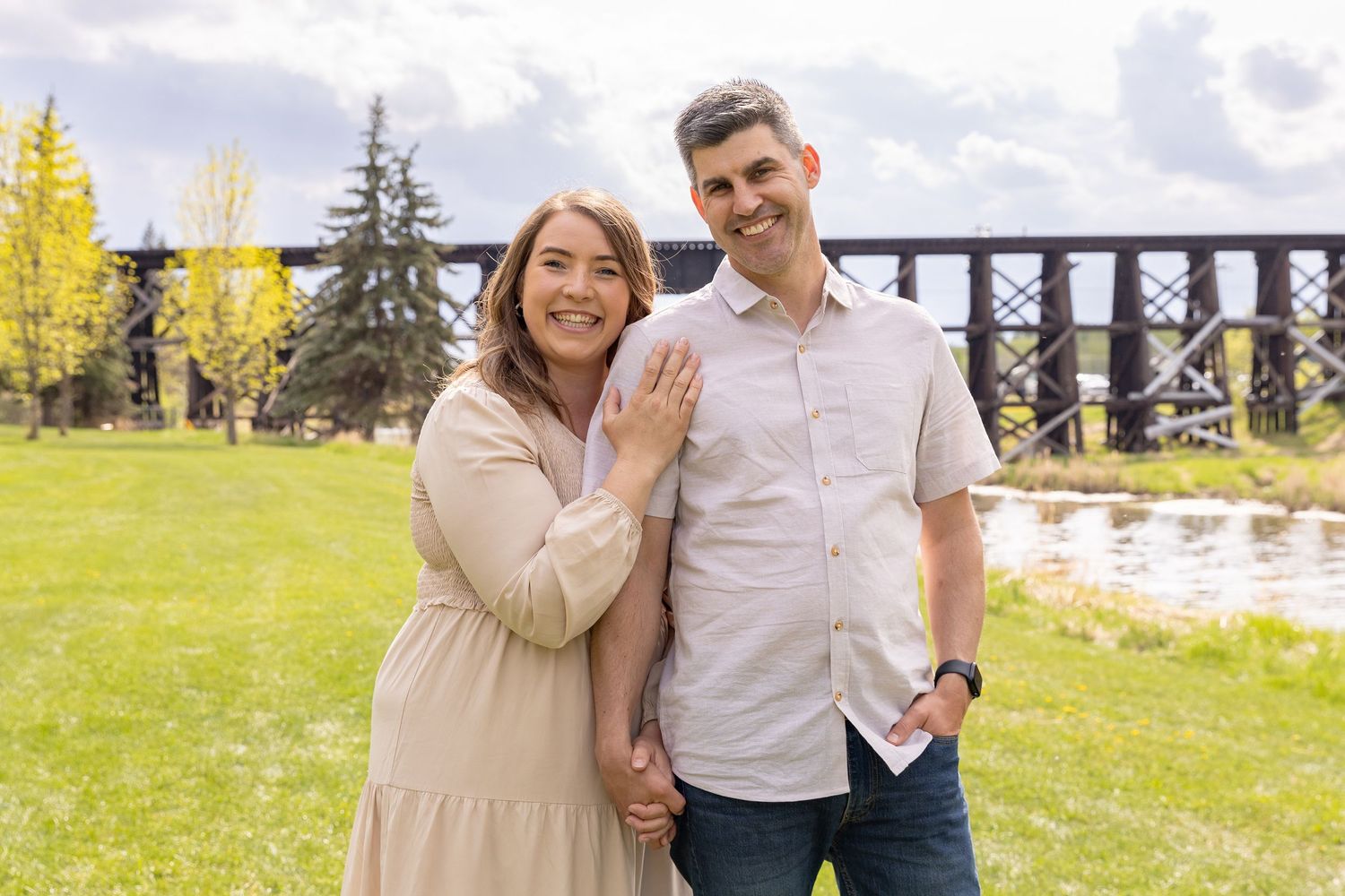 Smiling couple posing outdoors near a trestle bridge surrounded by green trees and grass.