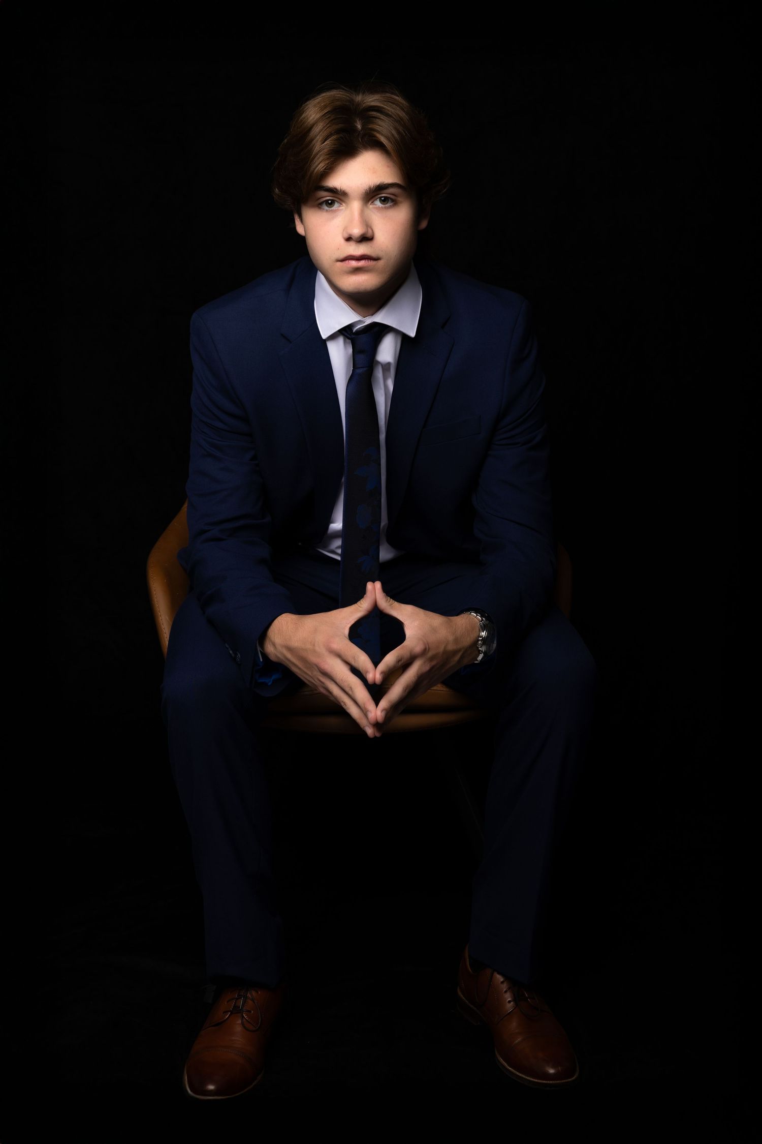 Confident young man in navy suit sitting with hands clasped against black background.