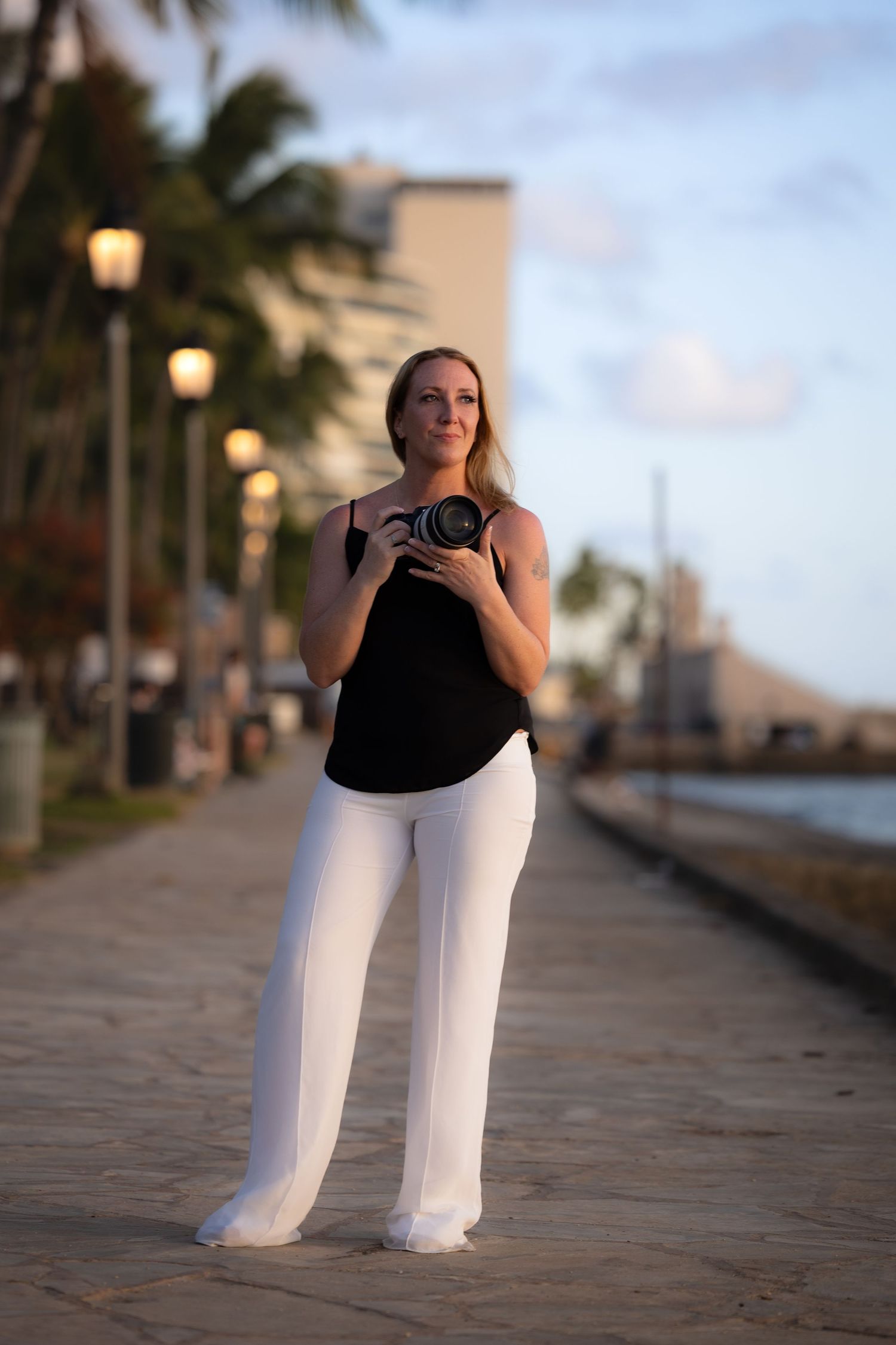 Female photographer holding camera standing on waterfront promenade at sunset with palm trees.