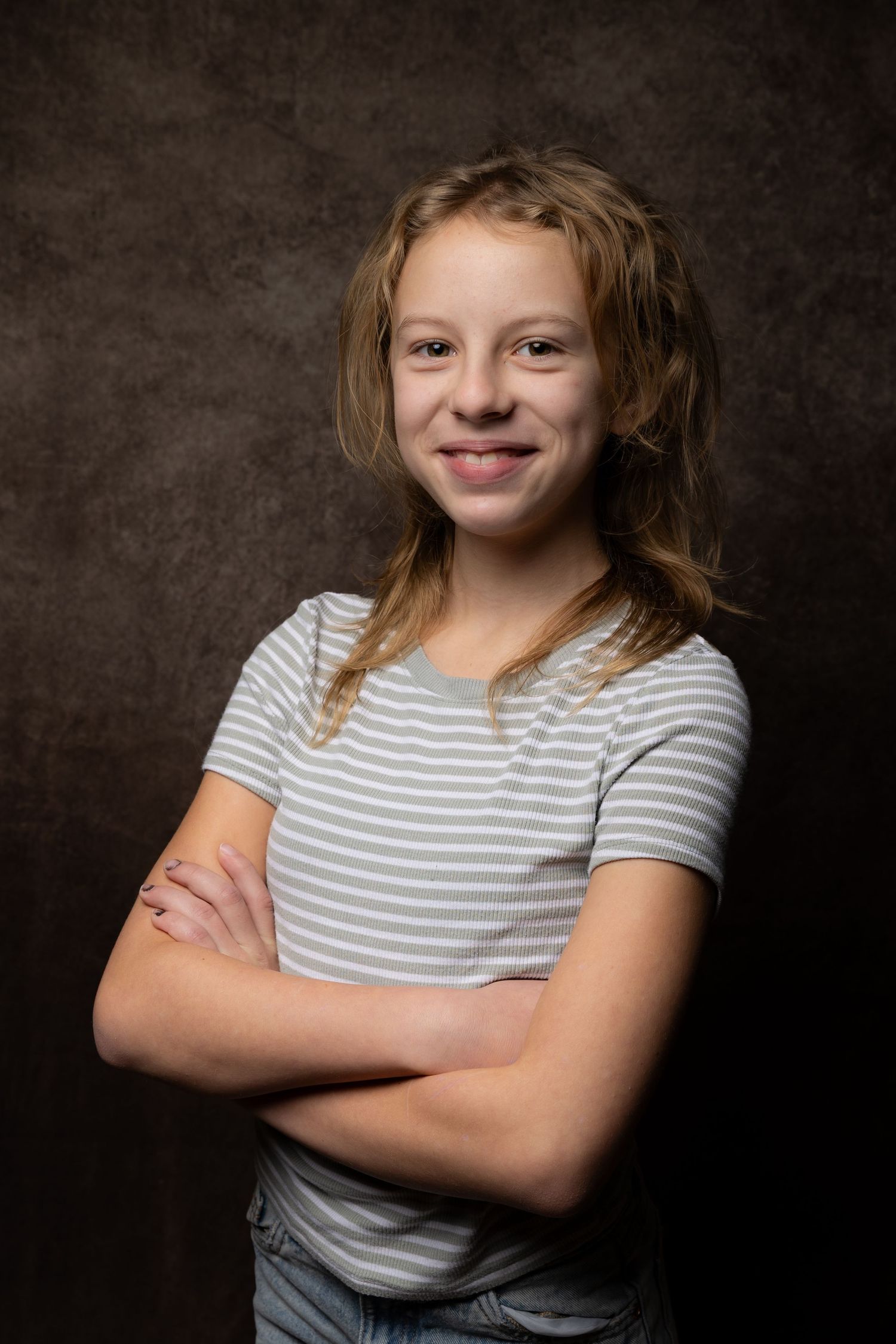 Smiling young girl with blonde hair wearing a striped t-shirt posing with arms crossed on dark background.