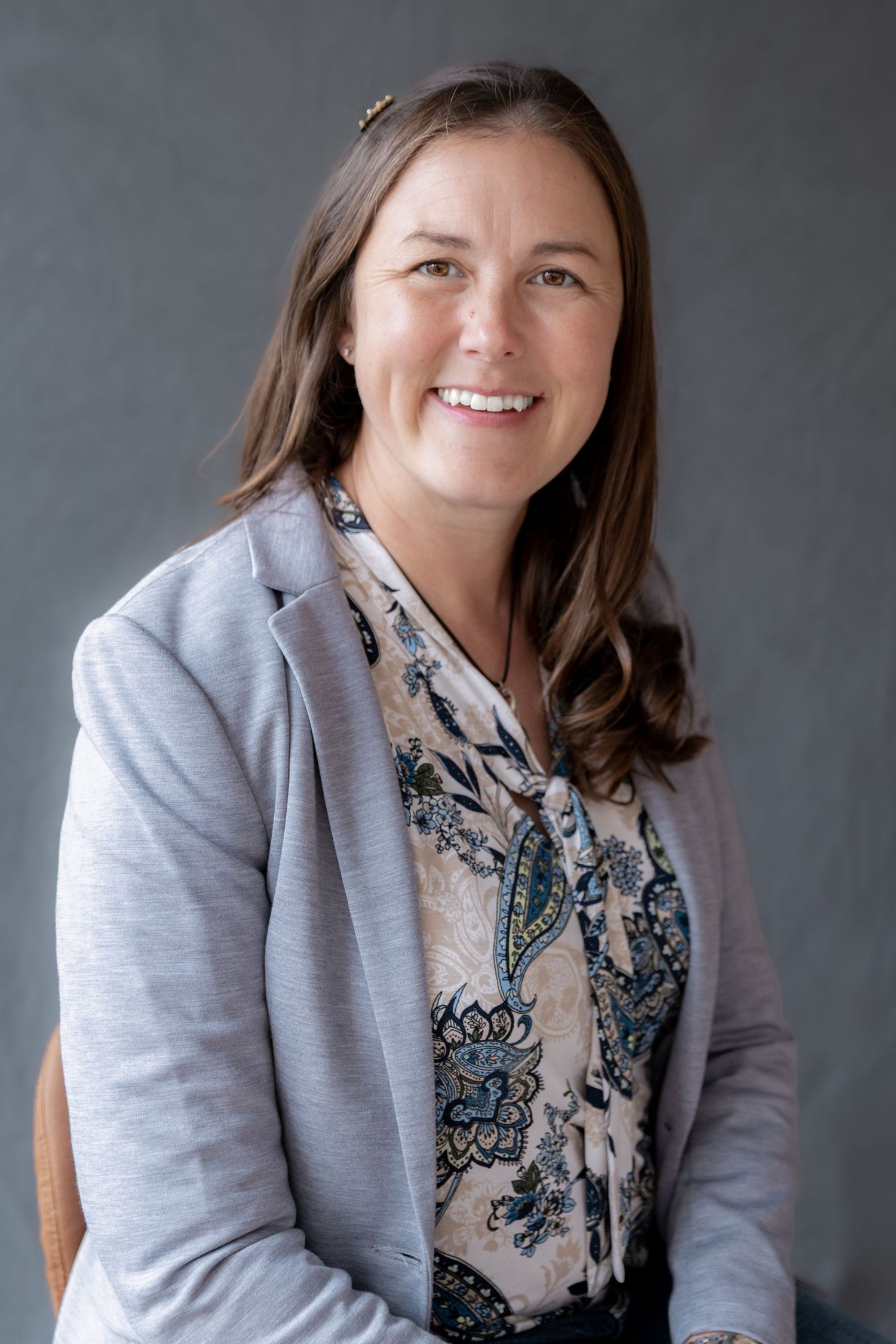 Smiling woman in a gray blazer and floral blouse seated against a gray background.