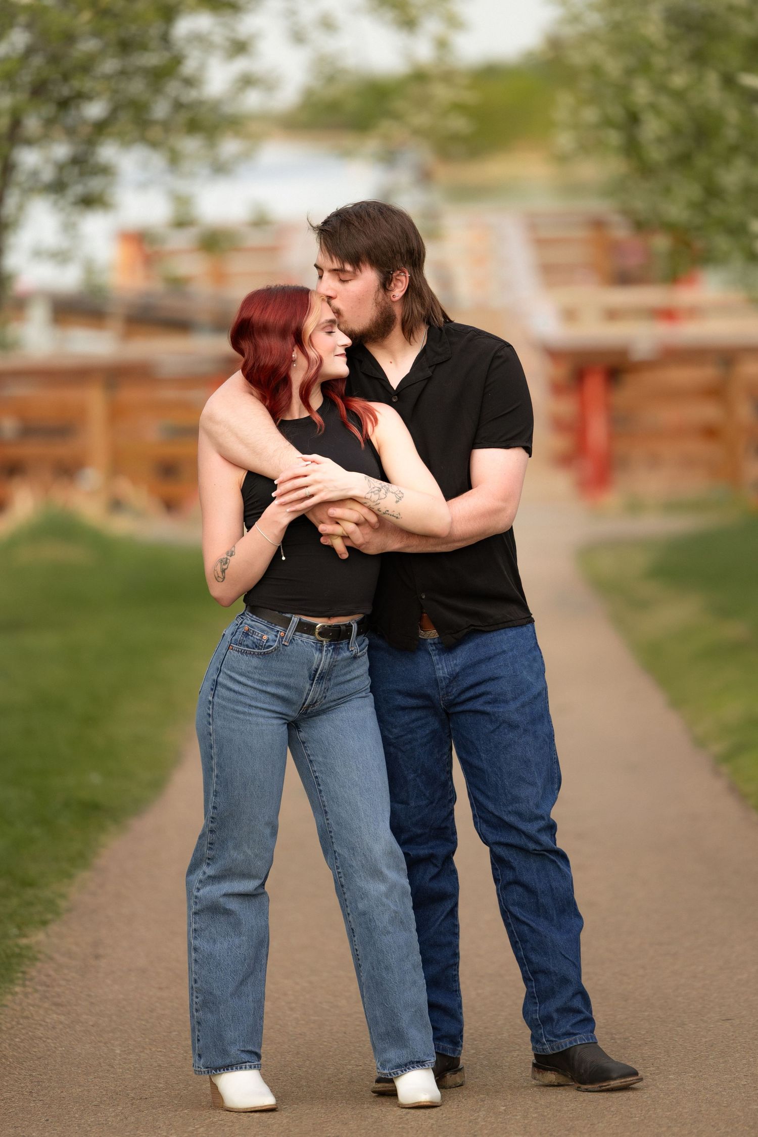 A couple in matching black tops and jeans share a romantic embrace on a park path surrounded by green trees.
