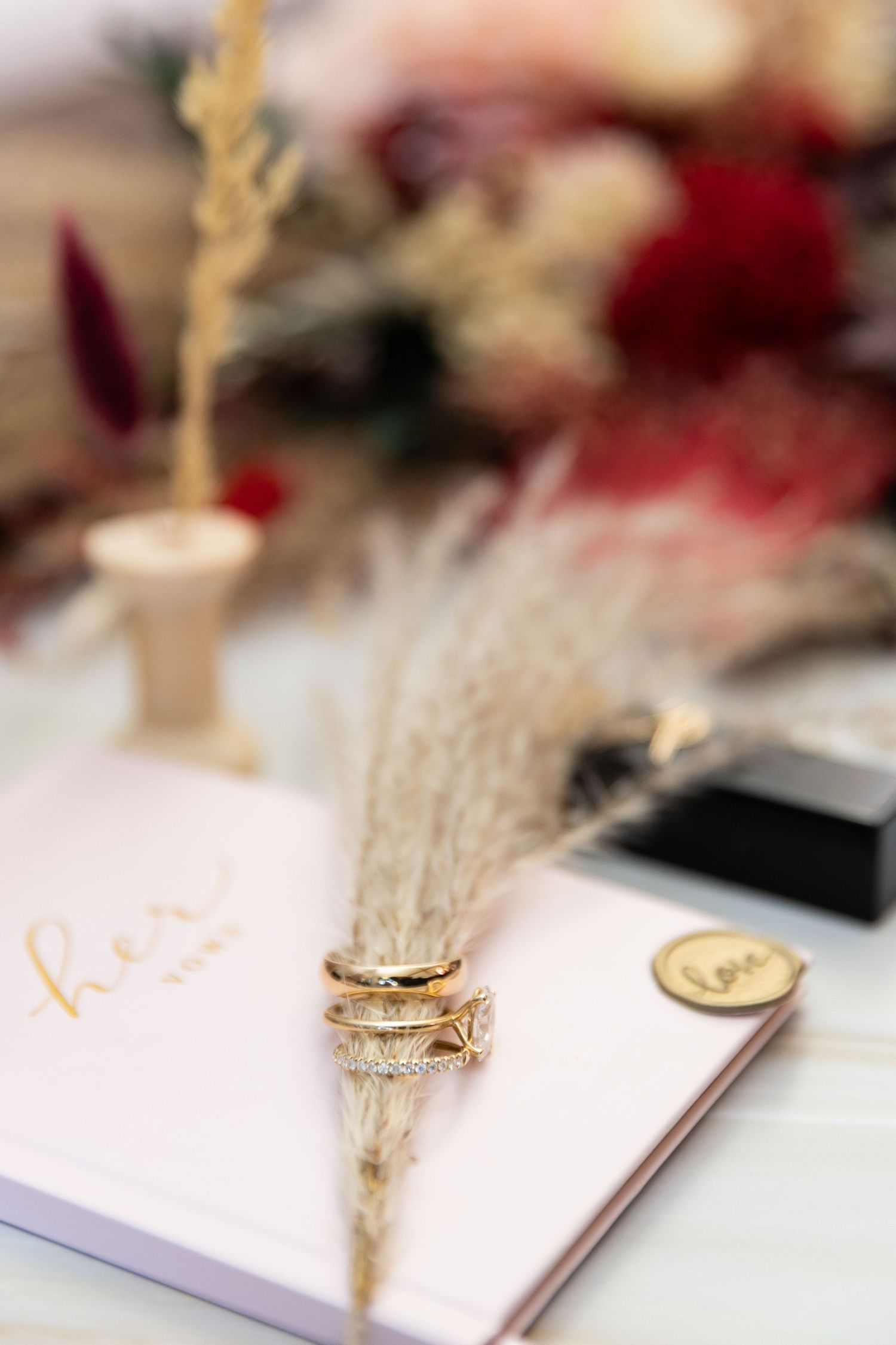 Gold wedding rings stacked on dried pampas grass atop a pink journal with floral decor in the background.