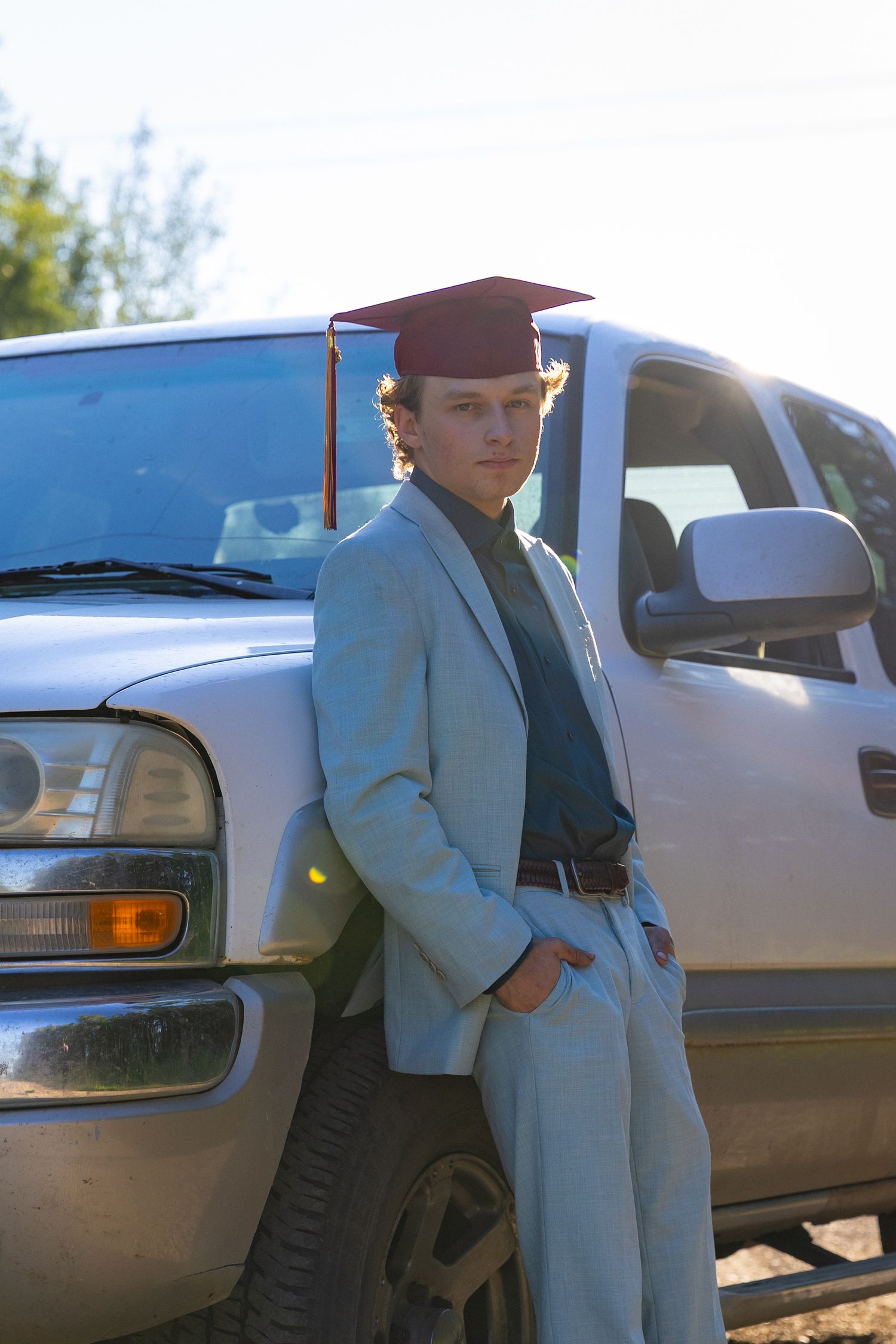 Graduating young man in a light blue suit and graduation cap leaning against a silver pickup truck outdoors.