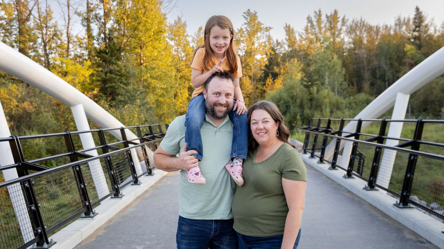 Happy family with a dad carrying daughter on shoulders and mom smiling on a white arch bridge.