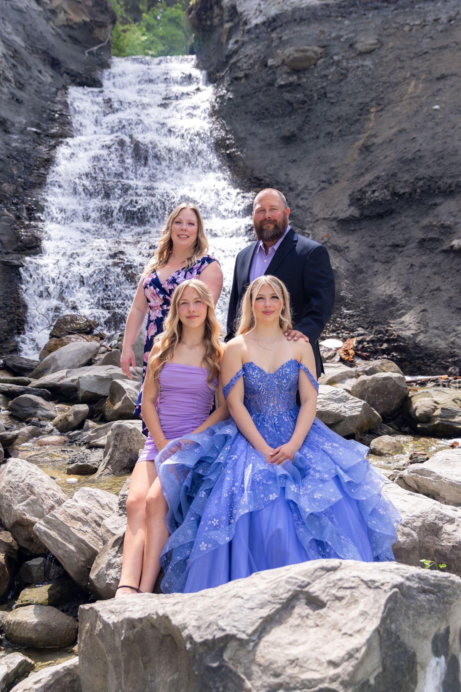 Family of four posing on rocks in front of a stunning waterfall, with two girls in purple gowns.