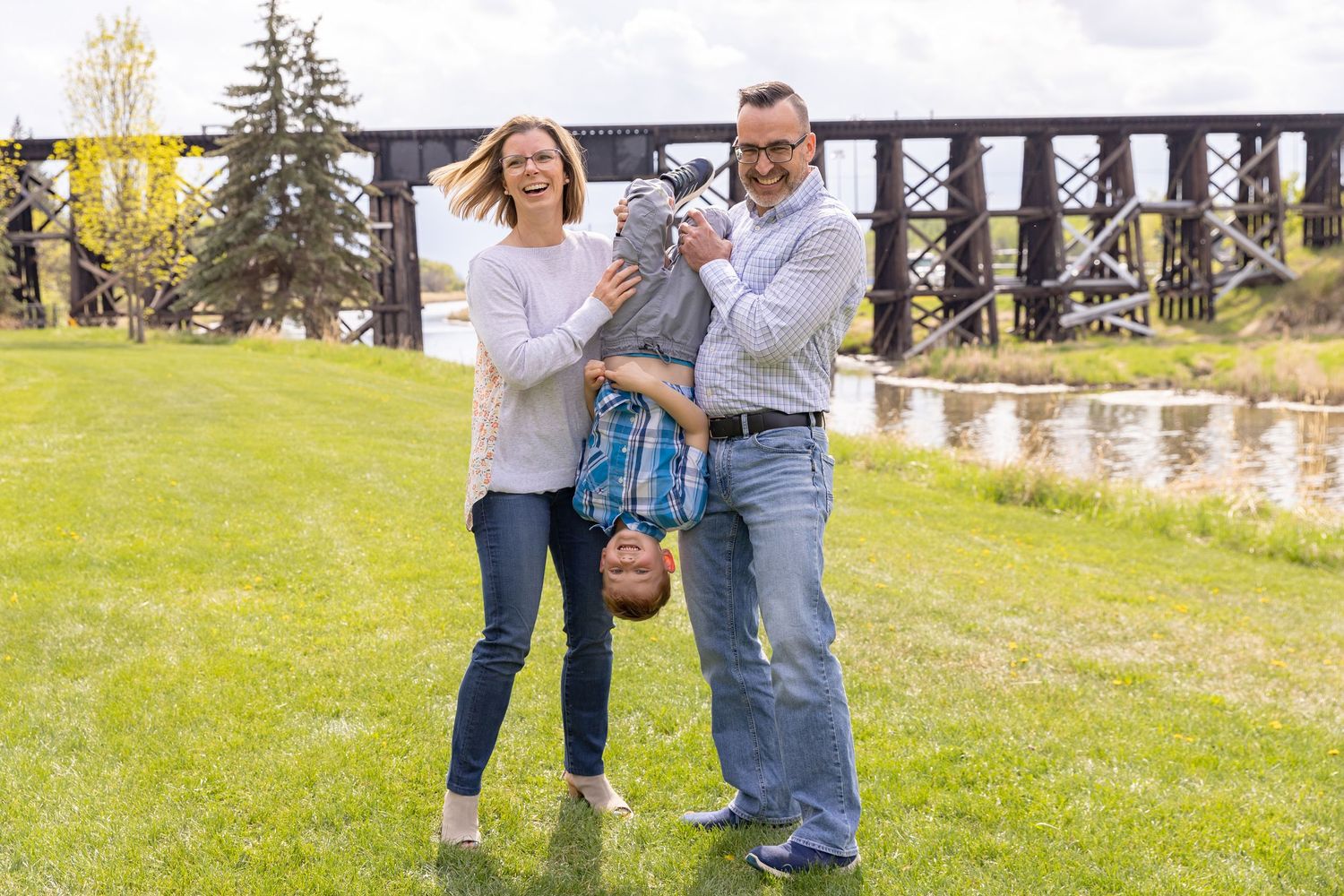 Happy family of three playfully holding child upside down in green park near historic bridge.