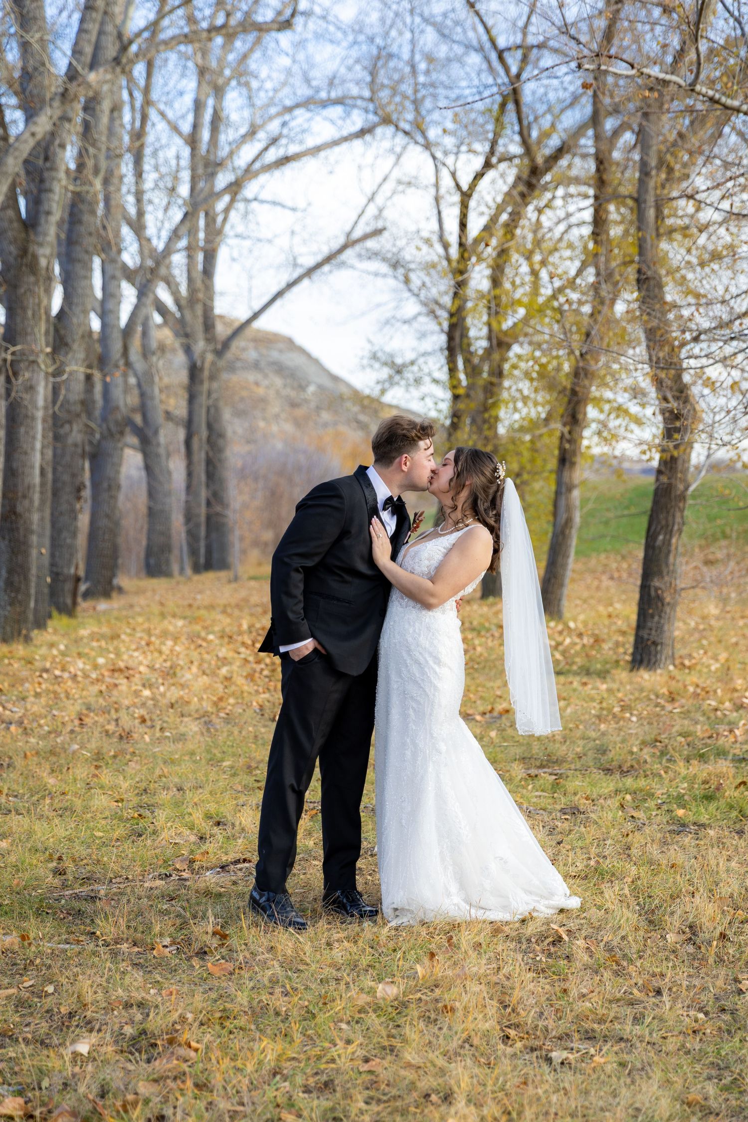 Newlywed couple sharing a kiss outdoors among bare autumn trees in wedding attire.