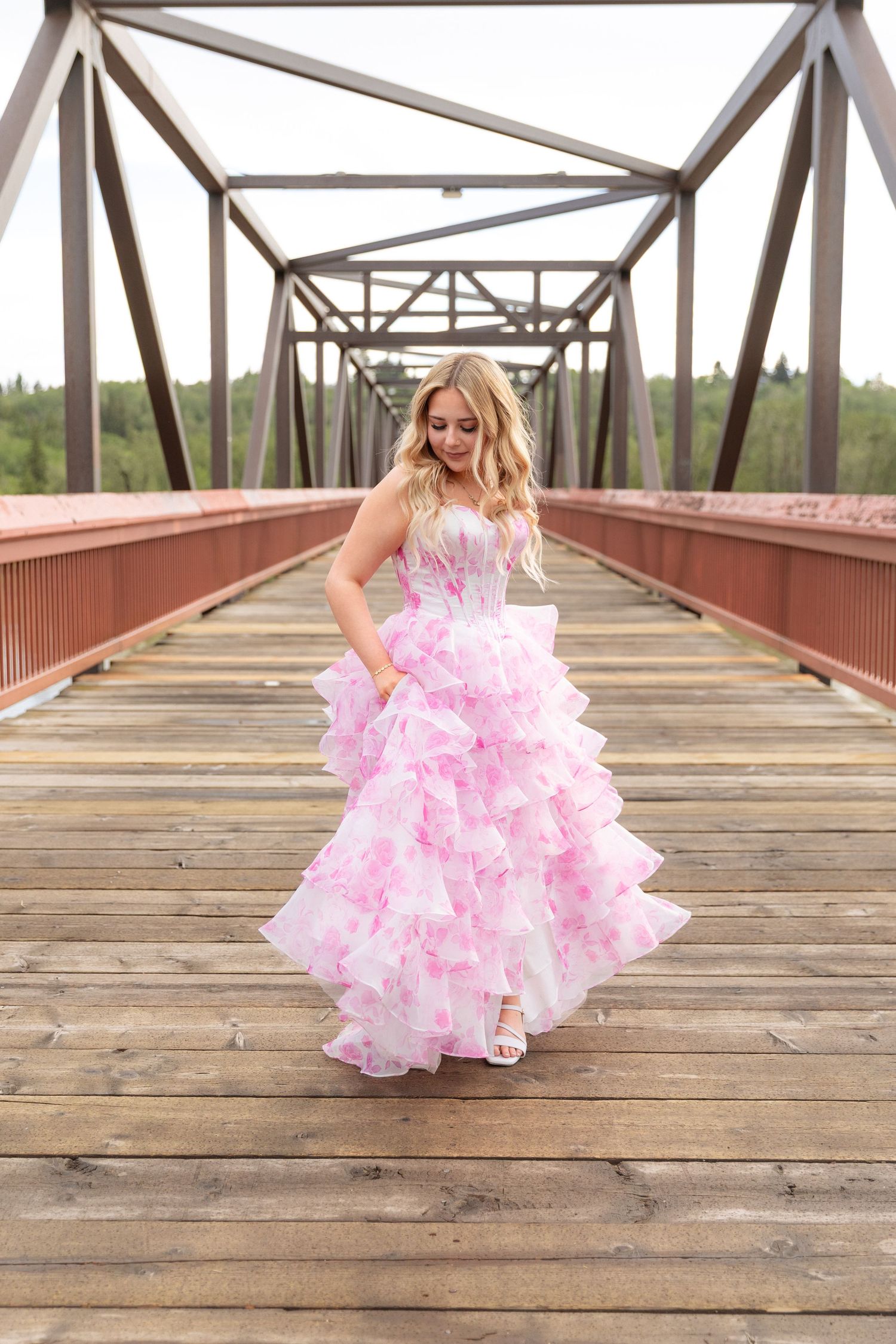 Graduating young woman in a pink floral ruffled ball gown twirling on a wooden bridge at golden hour.