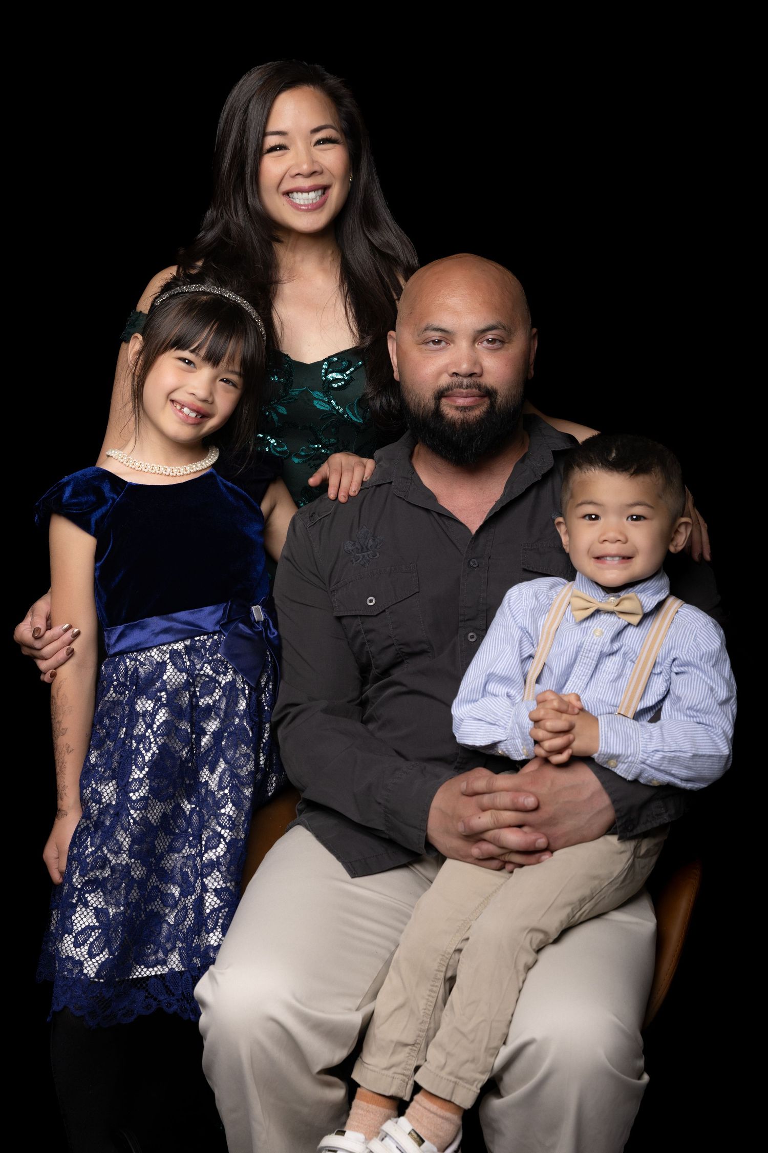 A smiling family of four poses together in formal attire against a black studio background for a portrait.
