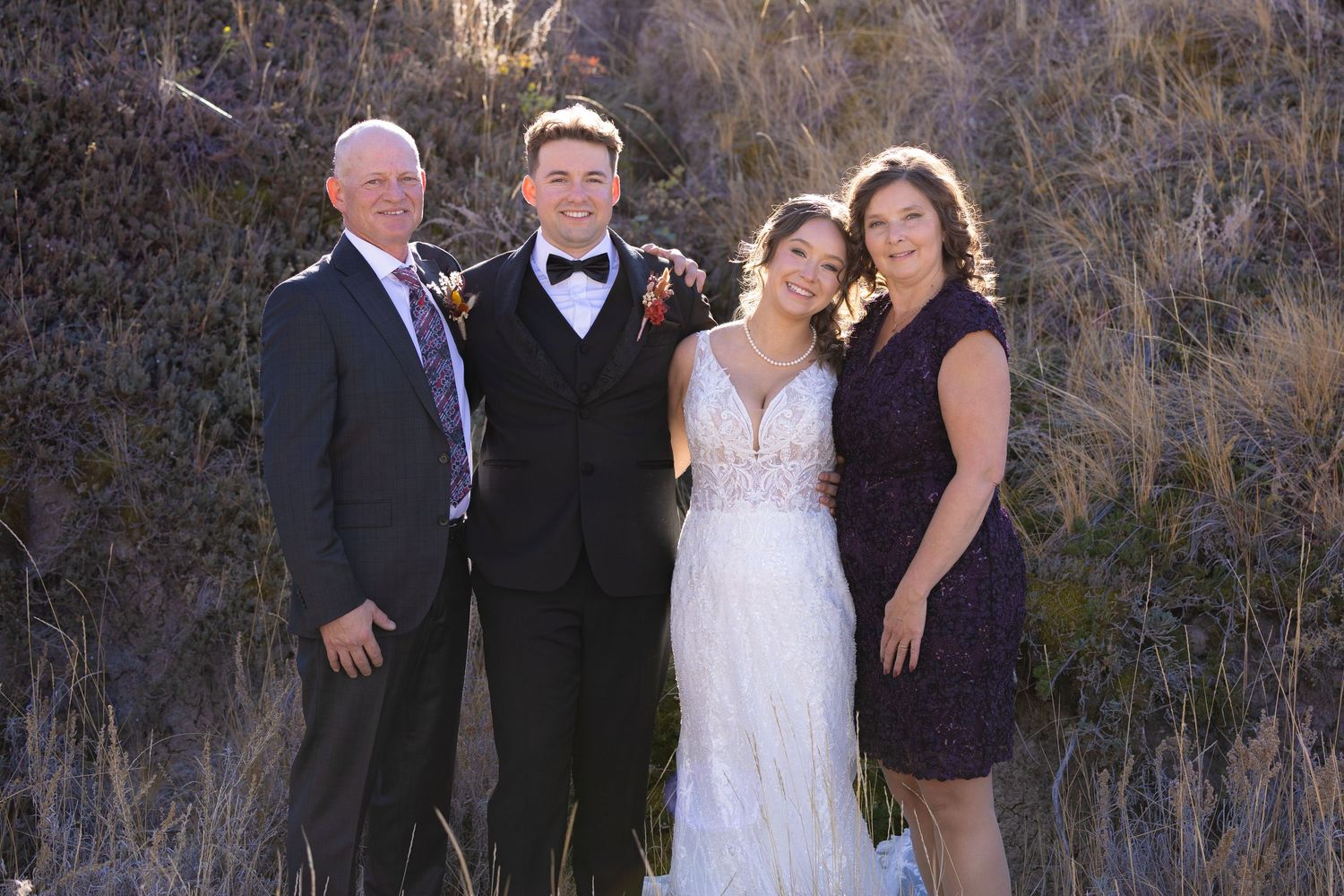 Wedding photo of bride and groom posing with parents outdoors in autumn landscape.