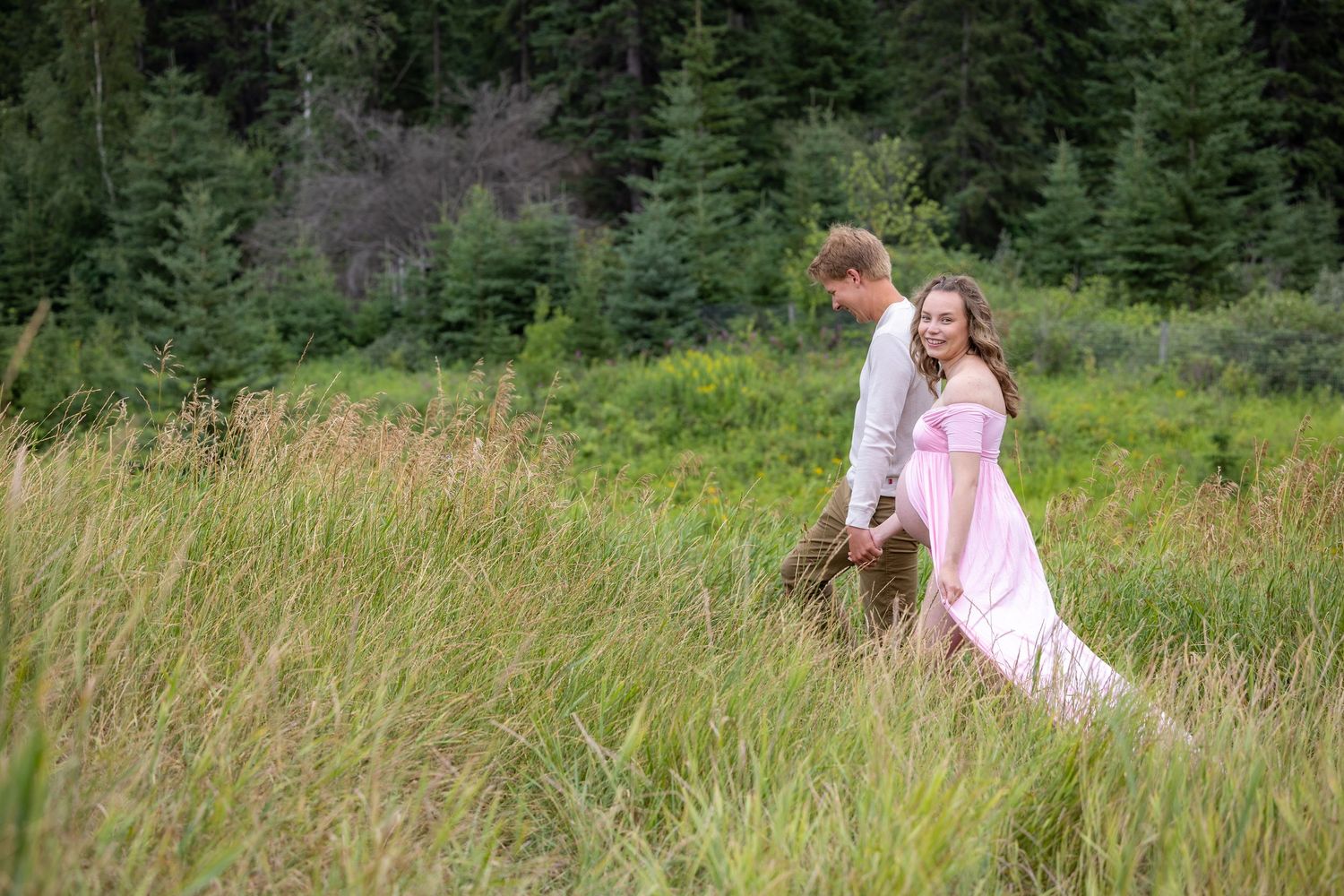 Couple walking through tall green meadow, woman in flowing pink dress, man in white shirt.