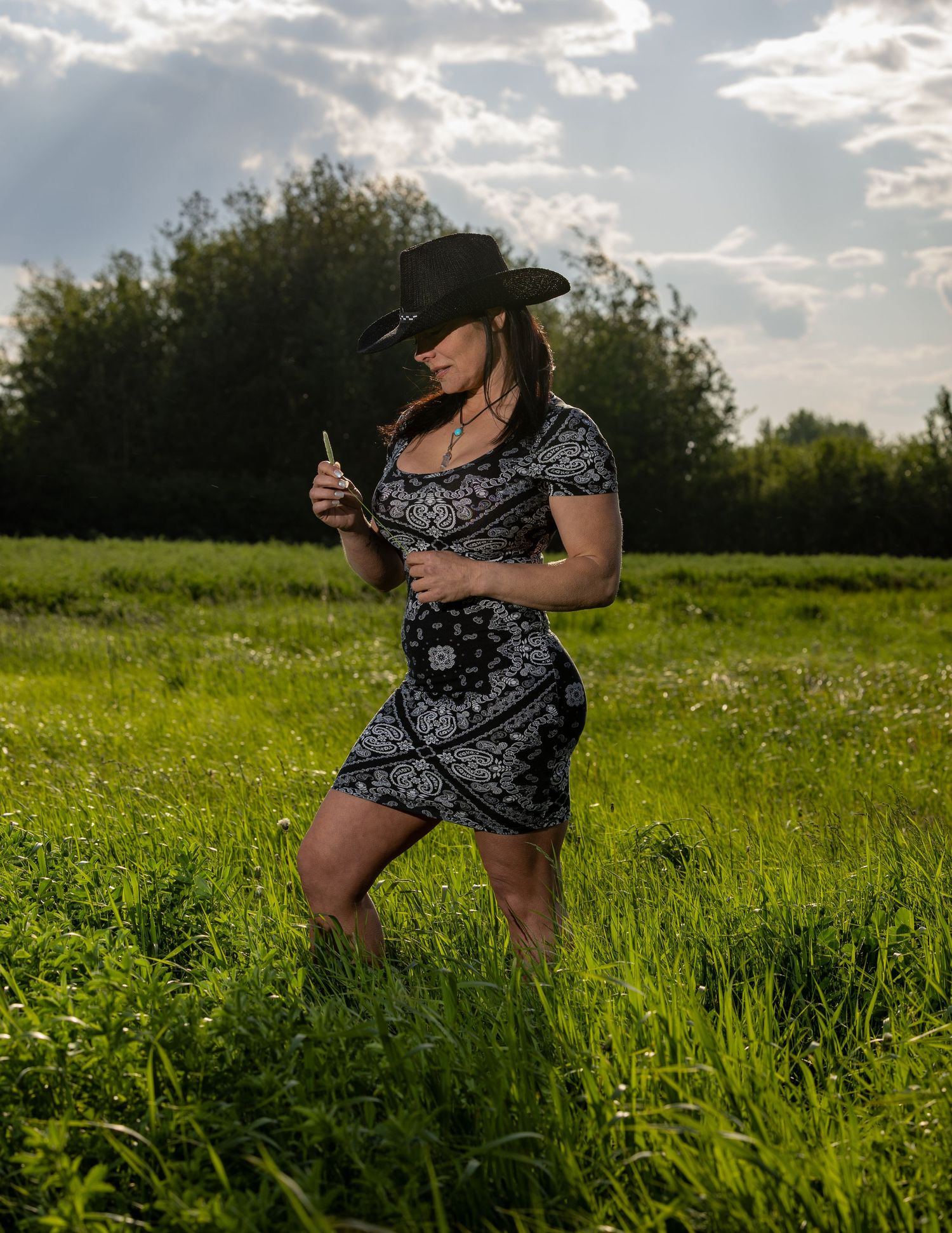 Woman in a black and white paisley dress and cowboy hat posing in a lush green field at sunset.