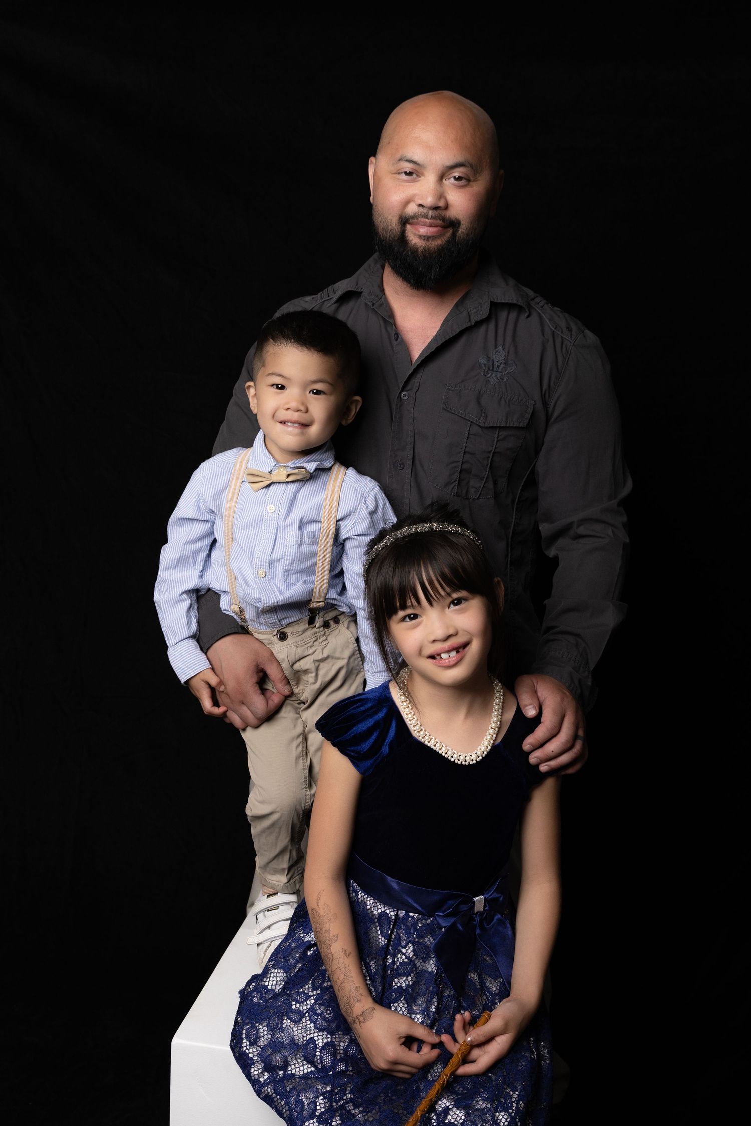 Father poses with young son and daughter in formal attire against black studio background.